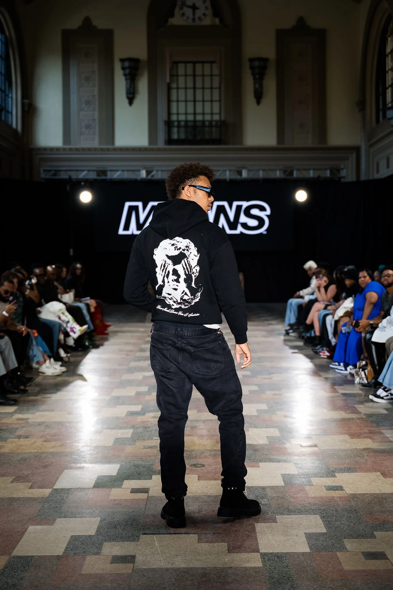 A male model on a runway, wearing a black hoodie with a graphic of a woman with hands covering her eyes on the back, black pants, and black boots. The setting appears to be an indoor fashion show with an audience and a large screen displaying the brand name in the background.