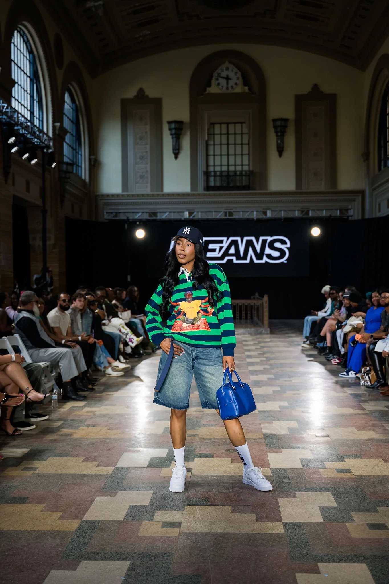 A fashion model walks down a runway in an indoor venue with an ornate ceiling and large arched windows. She wears a colorful oversized sweatshirt featuring a graphic of a person, baggy denim shorts, white sneakers, and a navy baseball cap with the Yankees logo. Audience members are seated along the sides of the runway.