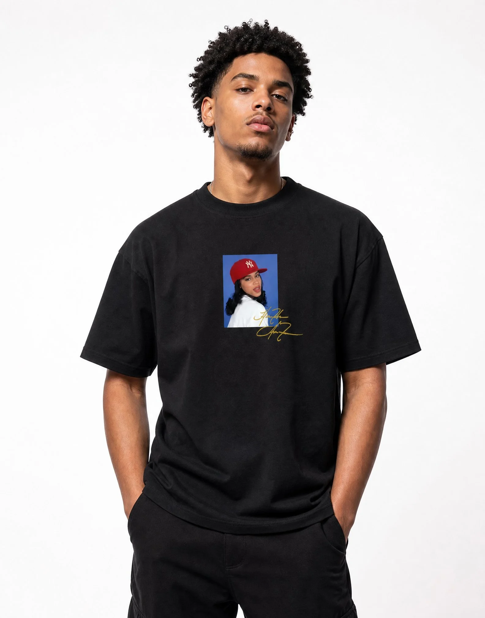 A young man with short curly hair and a light beard wearing a black T-shirt with a graphic of a woman in a red baseball cap on the front, standing against a plain white background.