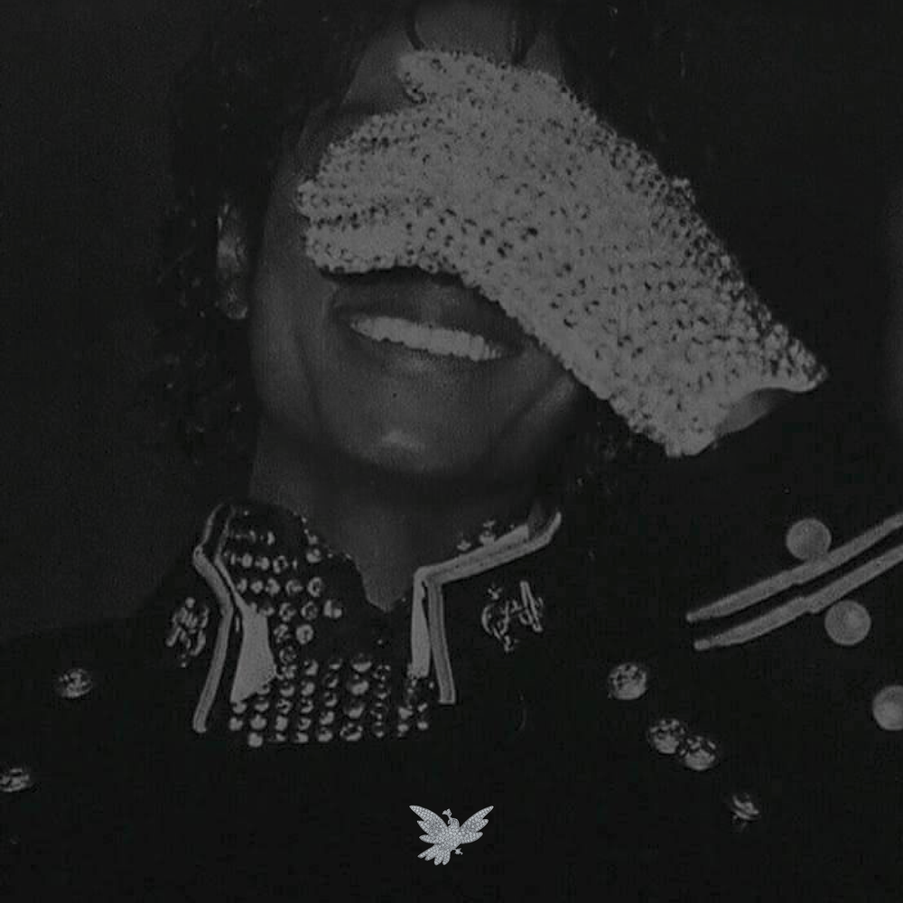 Black and white photo of Michael Jackson, smiling with a hands over her eyes, wearing a decorated jacket with beads and patches.