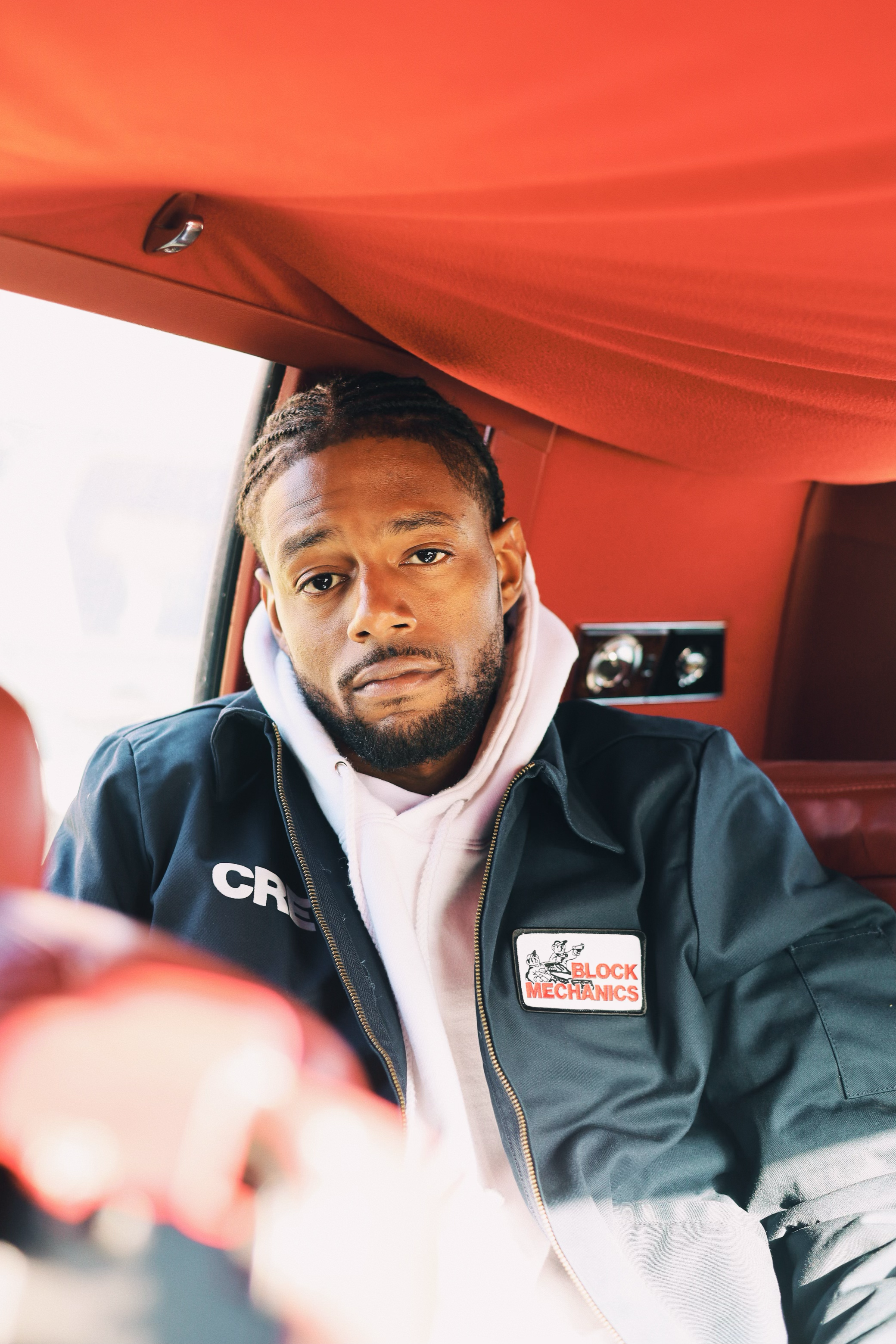 A man with short dreadlocks and a beard sitting inside a vehicle, wearing a black jacket with a "Block Mechanics" patch and a white hoodie underneath, looking at the camera.