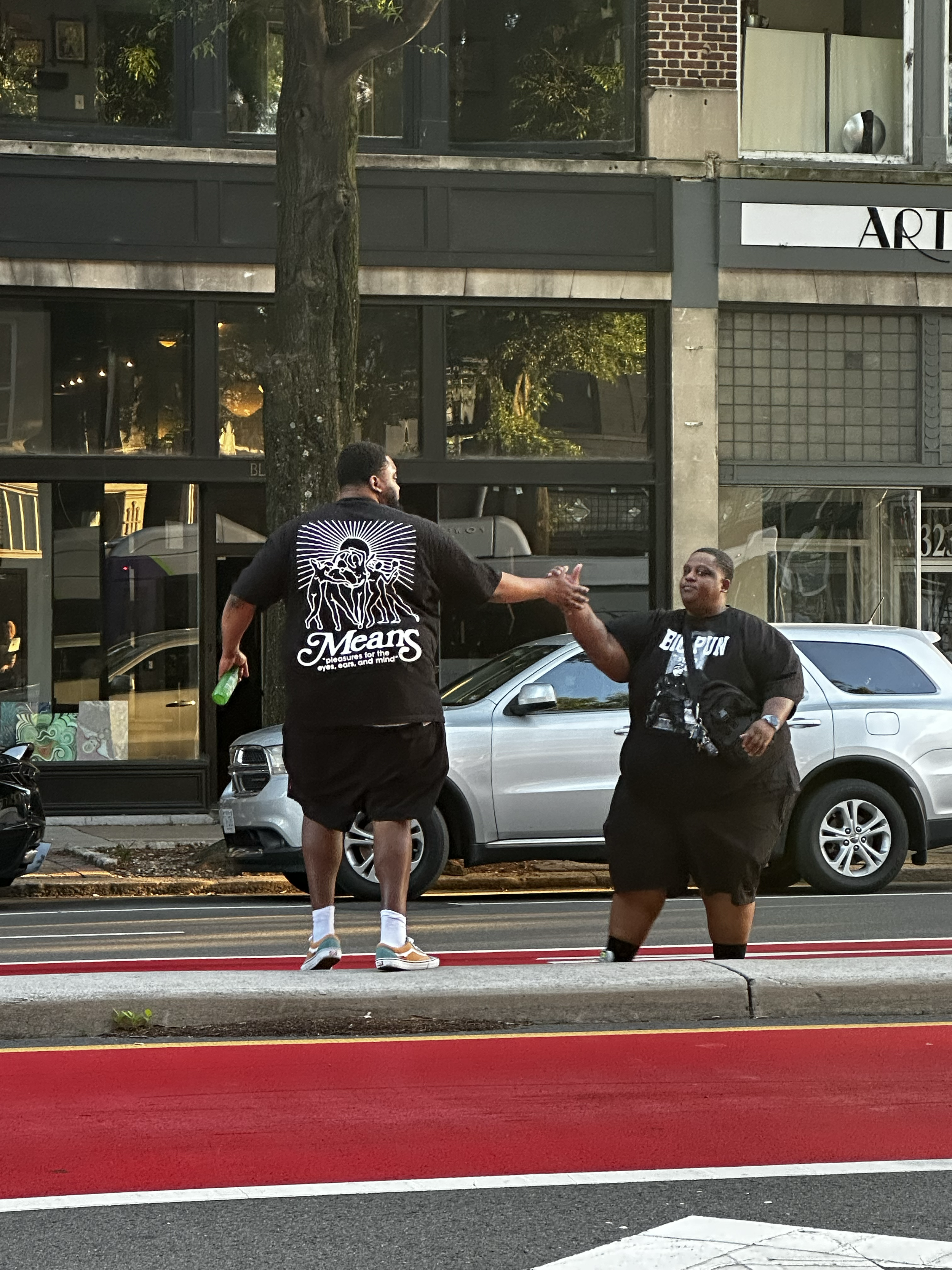 Two people standing on a city sidewalk, greeting each other with a handshake. One person is taller, wearing a black t-shirt with a design on the back, shorts, and sneakers. The other person is shorter, wearing a black t-shirt with white text and graphics, shorts, and carrying a crossbody bag. Cars are parked along the street, and a building with large windows is visible in the background.