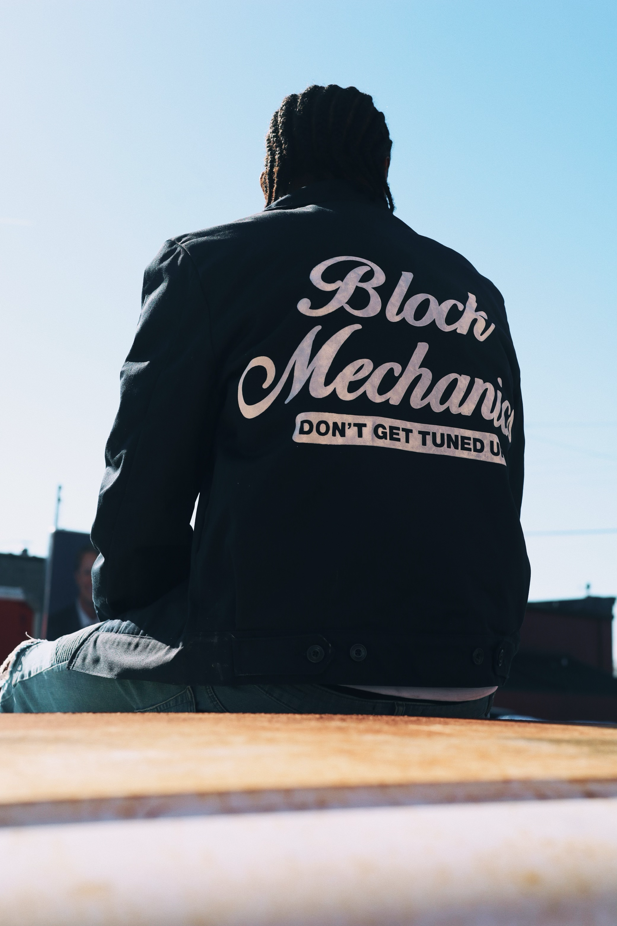 Person sitting outdoors on a wooden surface, viewed from the back, wearing a black jacket with white text that reads "Black Mechanic" and "Don't Get Tunned Up."