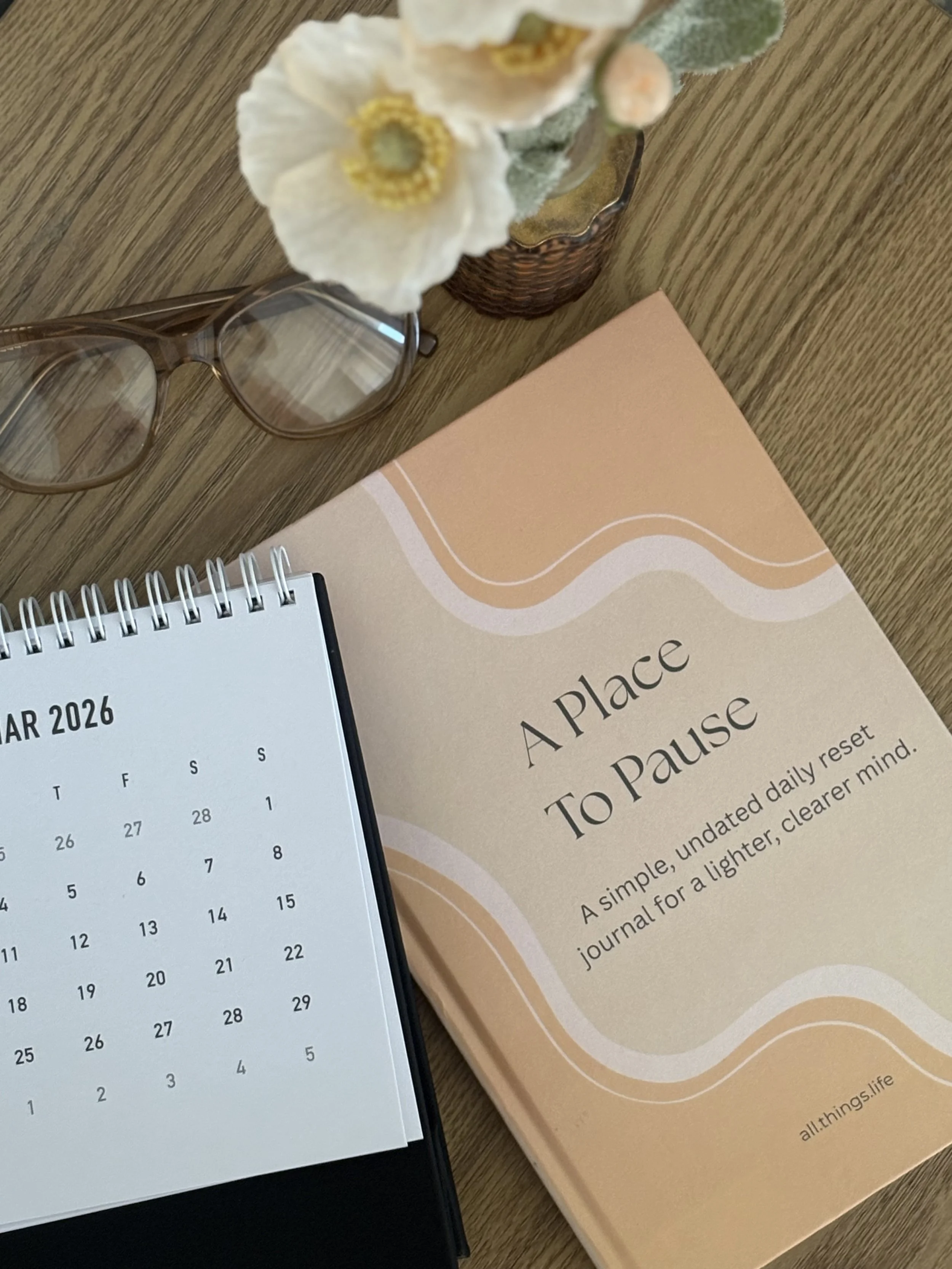 A flat lay of a desk with a pair of eyeglasses, a small calendar for March 2026, a spiral-bound notebook, a peach-colored journal titled "A Place To Pause," a small decorative flower arrangement in a wicker basket, and a closed notebook.