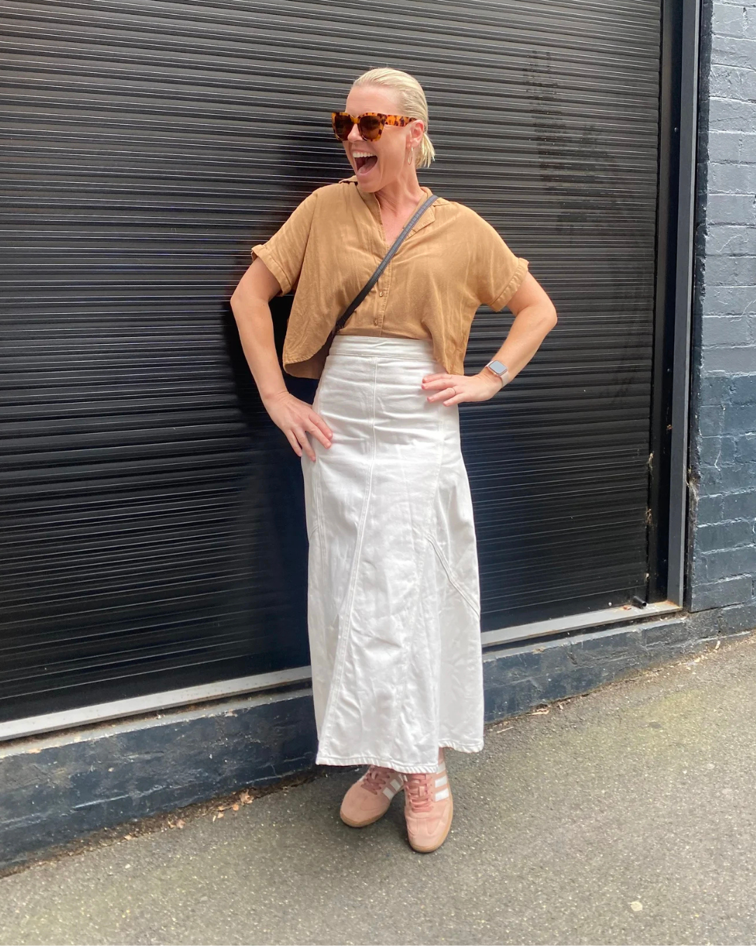 A woman with blonde hair in a ponytail, wearing large tortoiseshell sunglasses, a tan loose button-up shirt, a long white skirt, pink sneakers, and a smartwatch, standing outdoors in front of a black metal door and gray brick wall, smiling and laughing.