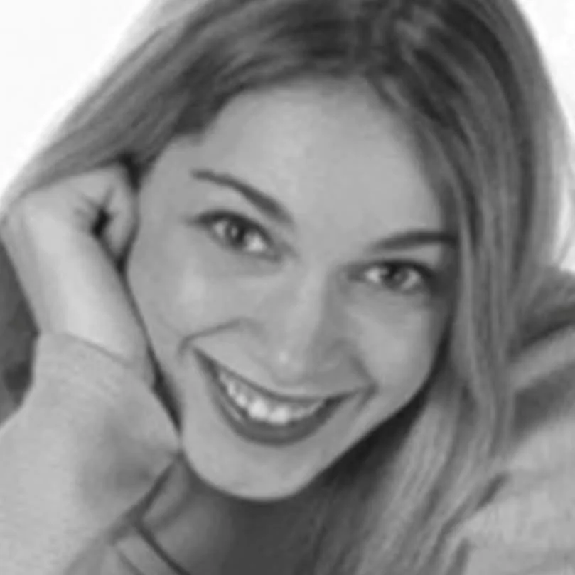 Black and white close-up of a Gina with long hair. They rest their hand on their cheek, exuding warmth and friendliness.