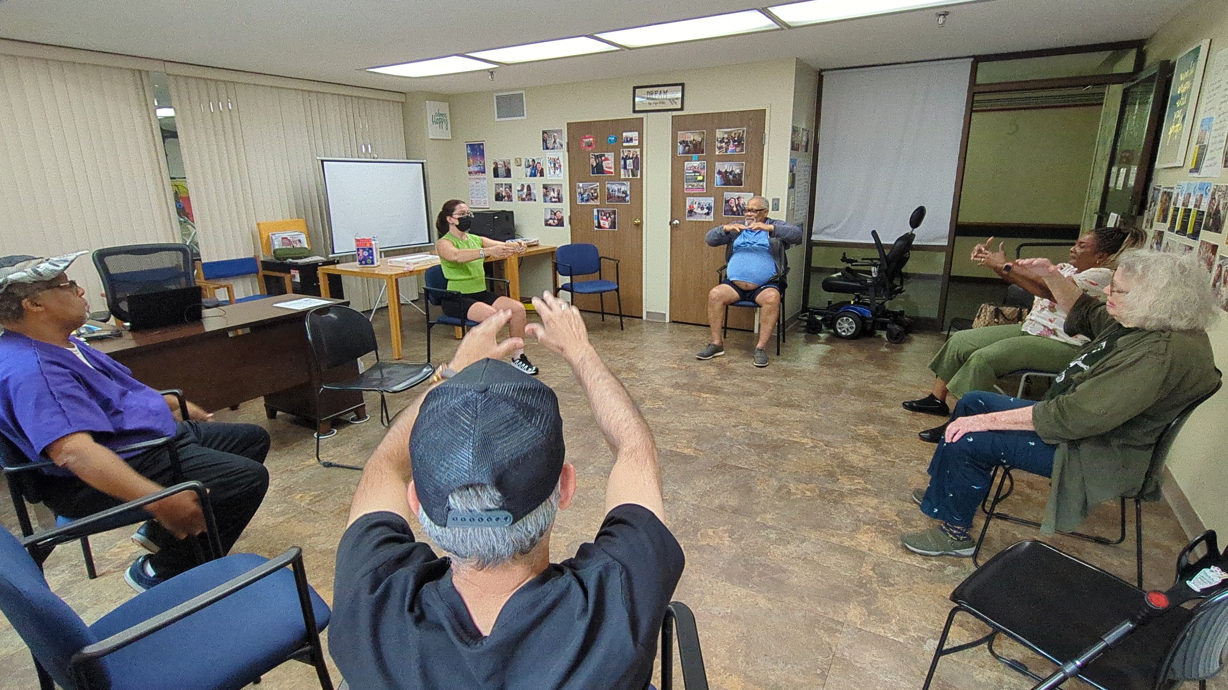  Teacher Madeline guiding a class in an elder center. All seated in a circle with arms raised.  