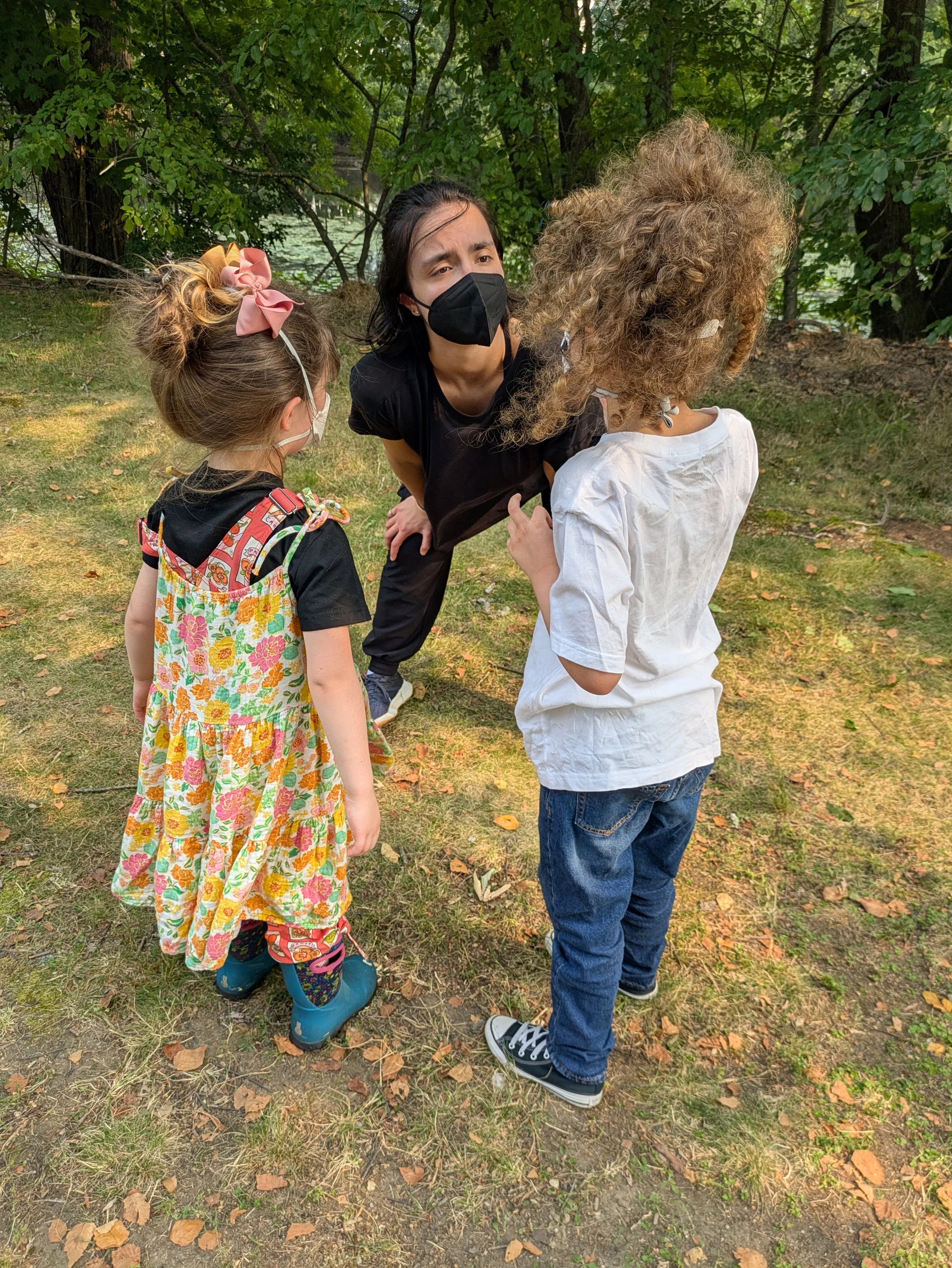 Teacher Ariana bending down to talk to two young dance students. All masked in an outdoor setting.  