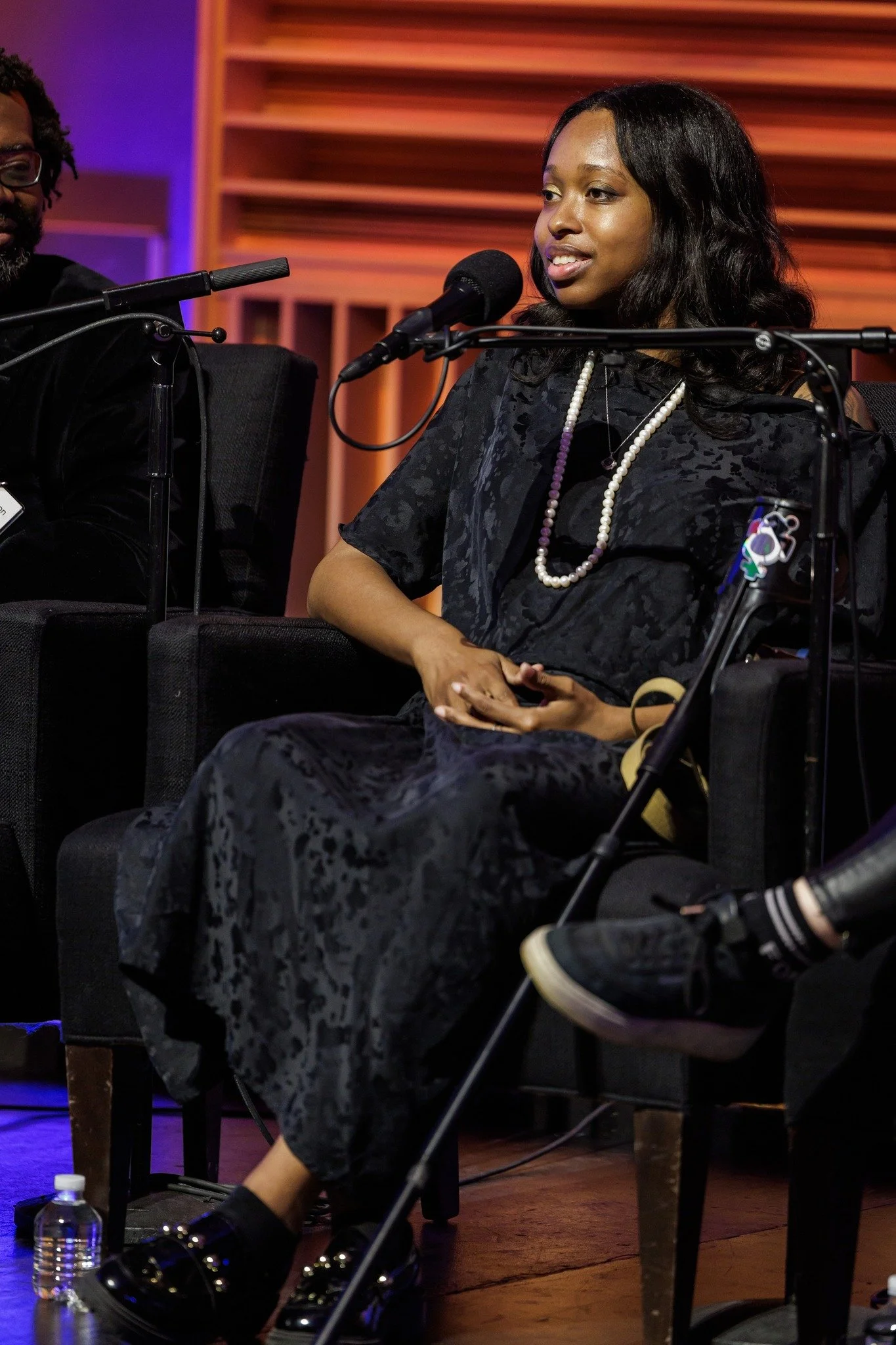  Ellice in a black dress and pearls, seated with a microphone at mouth, speaking on a panel. 
