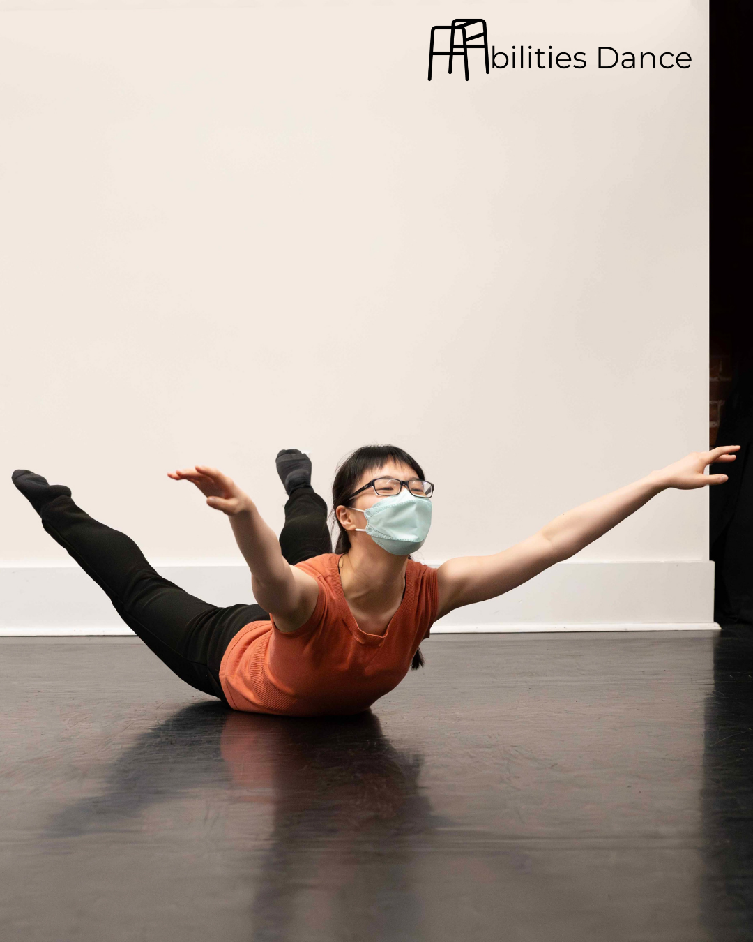 Linda in an orange top and black leggings. On ground with legs and arms raised up as she looks up. in a starfish type pose.