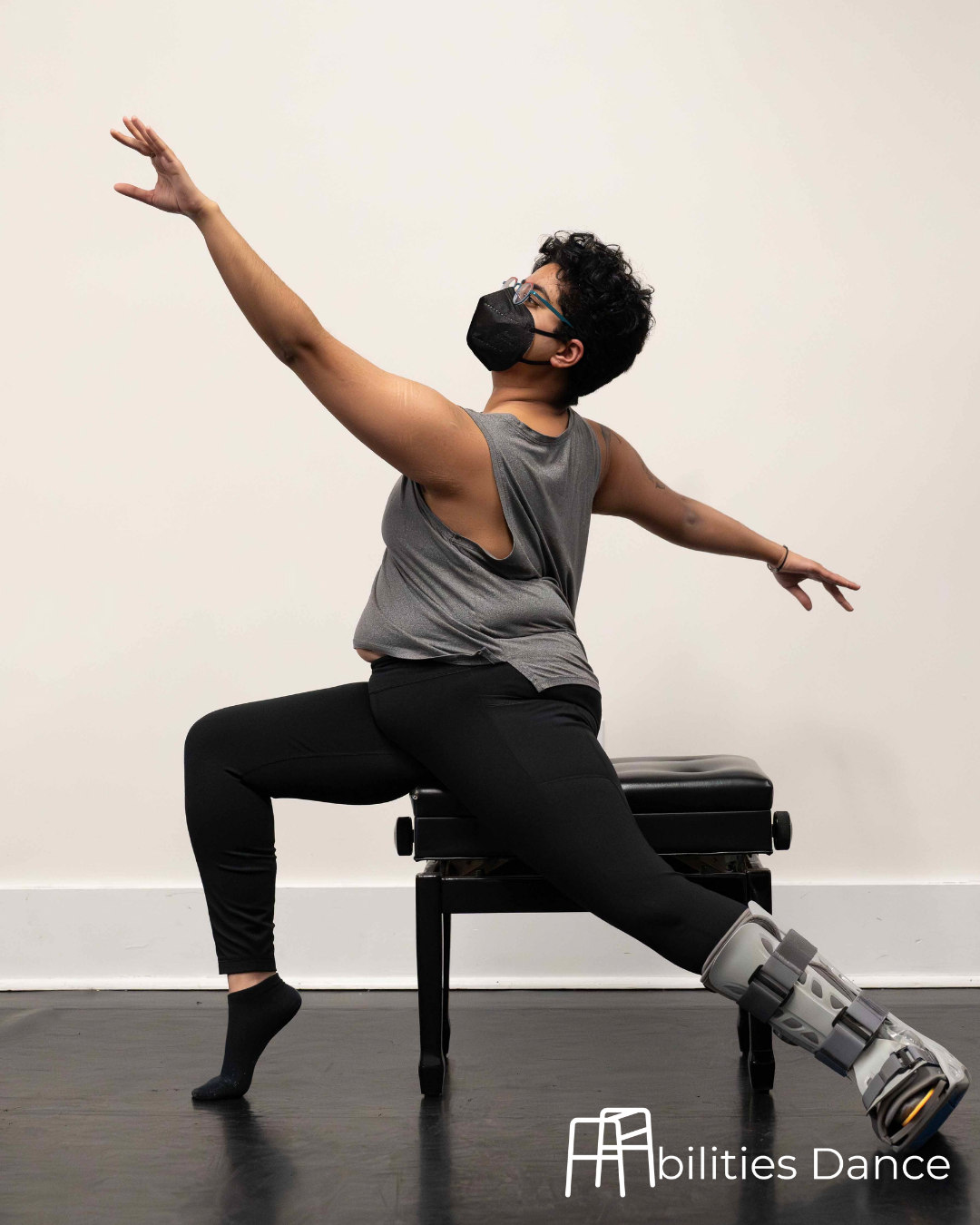 Image of Amaal in a grey top and leggings. Seated on a stool with arms on a diagonal looking up. One foot pointed forward. The other extended with a footbrace on