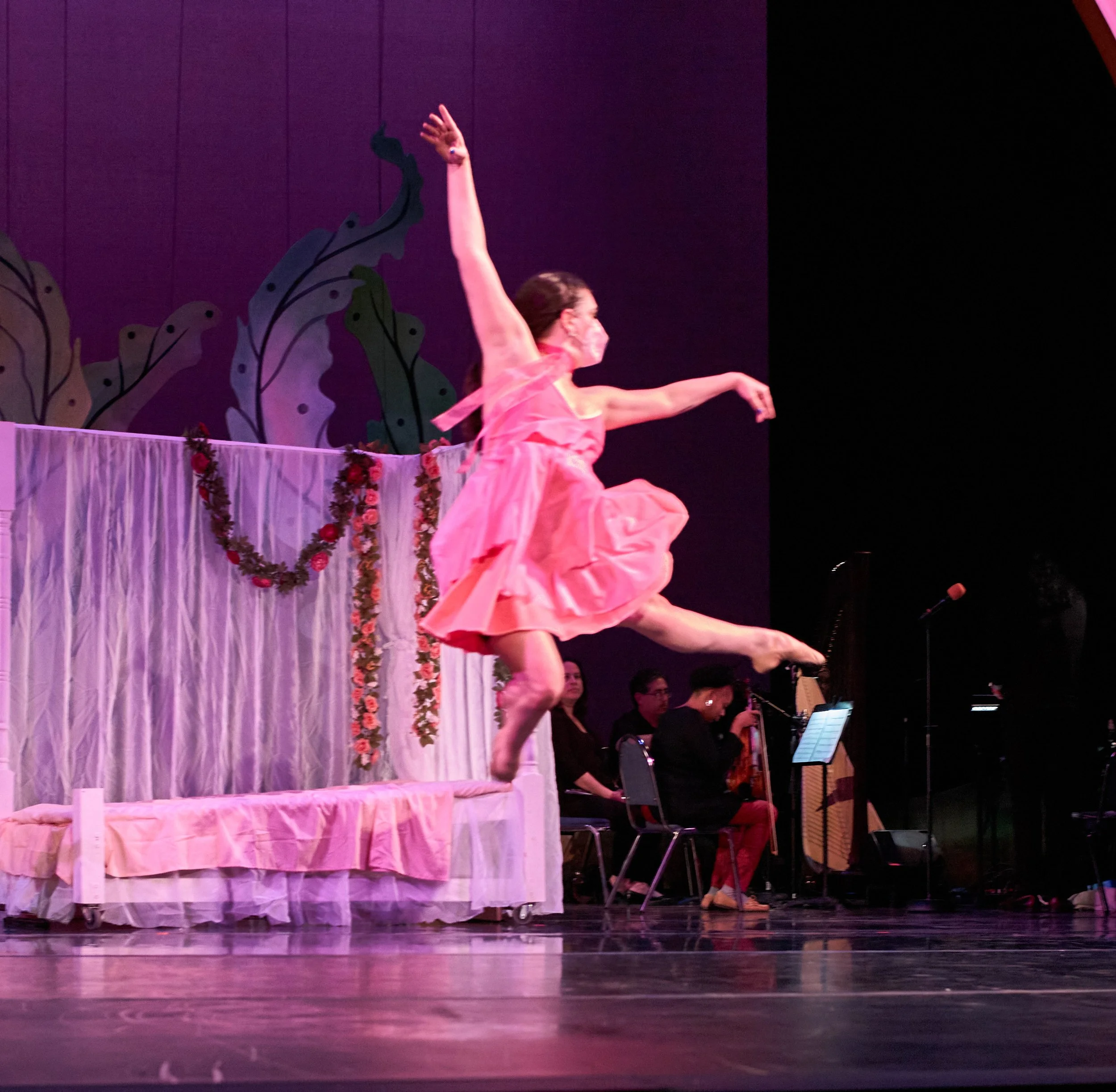 Image of Dara mid leap with one leg bent in and one leg extended with arms up.. She is wearing pink dress and mask as Sleeping Beauty. Behind her is a bed with a canopy of sheer fabric and flower chains.