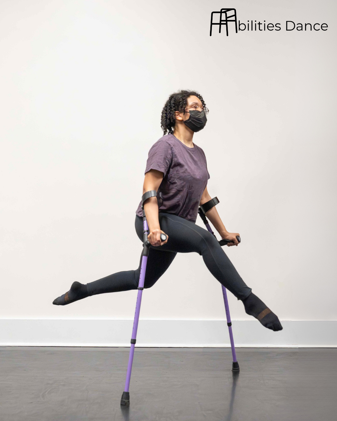 Image of Blake mid jump with legs bent while balancing on crutches. Wearing purple top with head faced forward.
