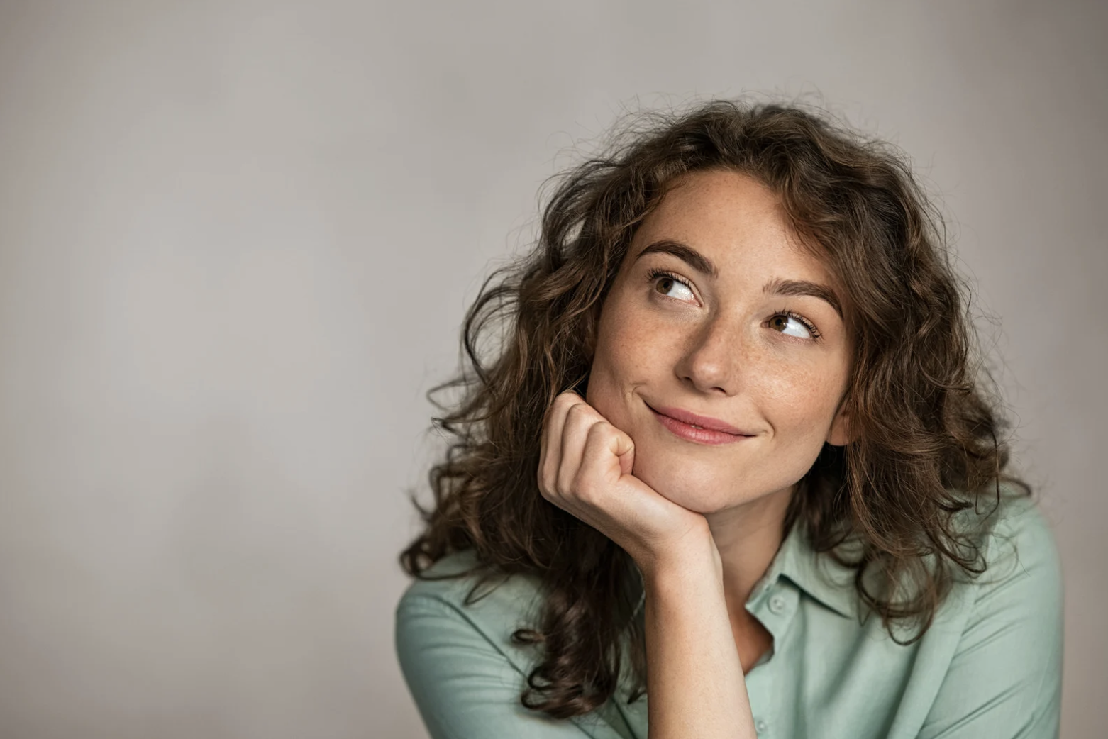 Woman with curly brown hair resting her chin on her hand and looking upward thoughtfully against a neutral background.