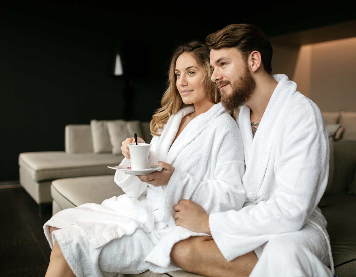 A couple wearing white spa robes relaxes together with a warm drink, enjoying a quiet moment in a luxurious, serene setting.