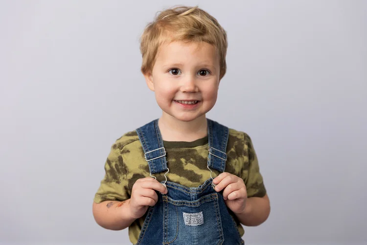 Child smiling in overalls against a light gray backdrop