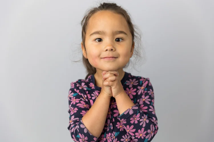 Child smiling with hands together against a light gray backdrop