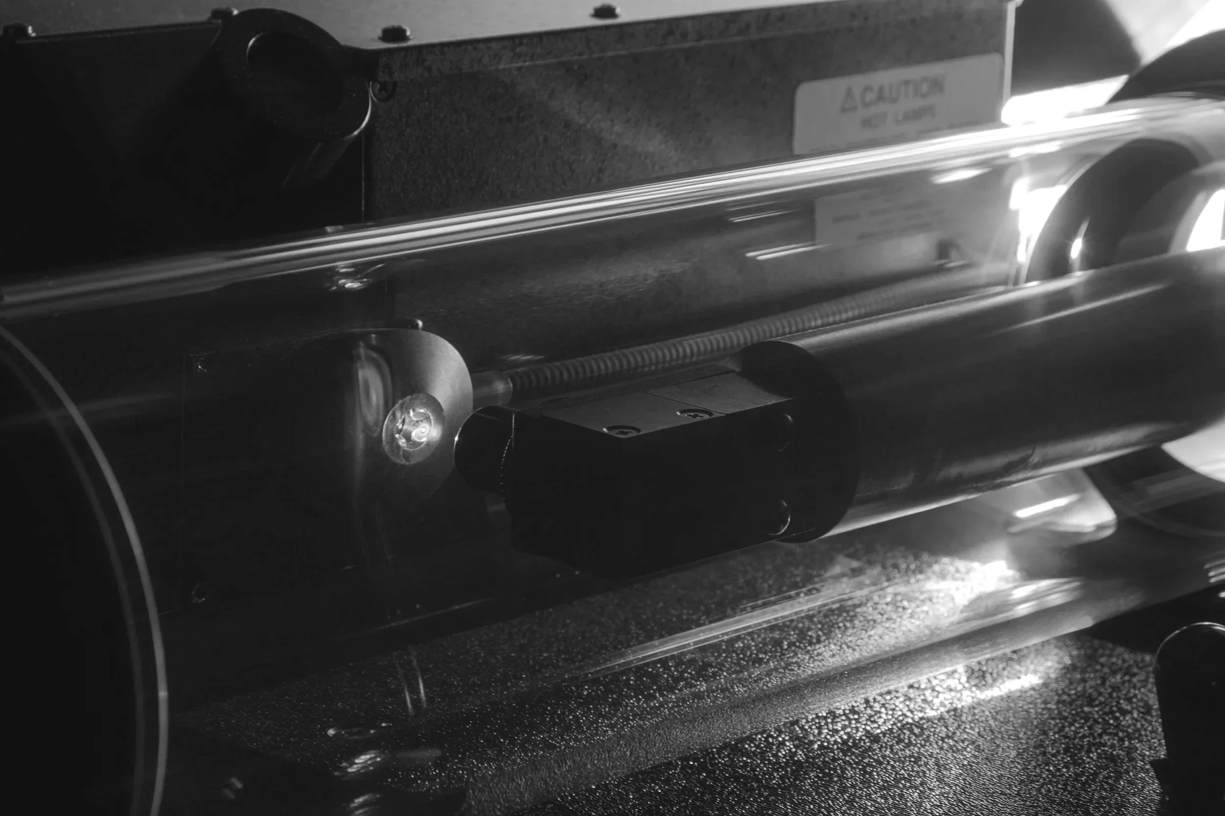 A close-up black and white photo of a mechanical device with a caution label, metal rollers, and a conveyor or track system.