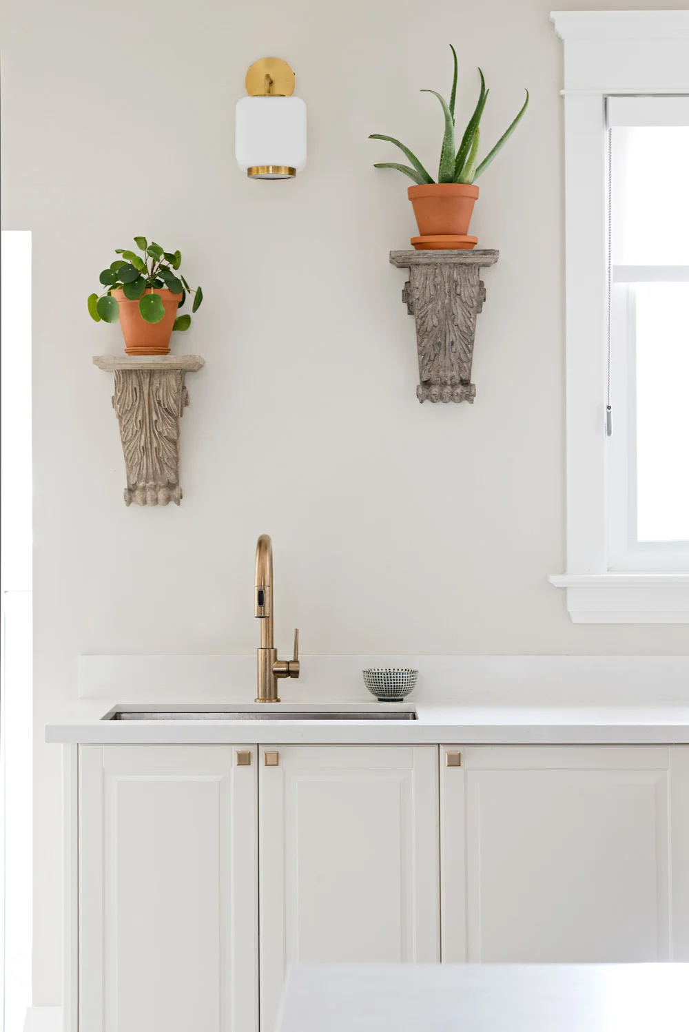Staggered wall mounted corbels with plants above kitchen sink with brass faucet, cabinet knobs and sconces.