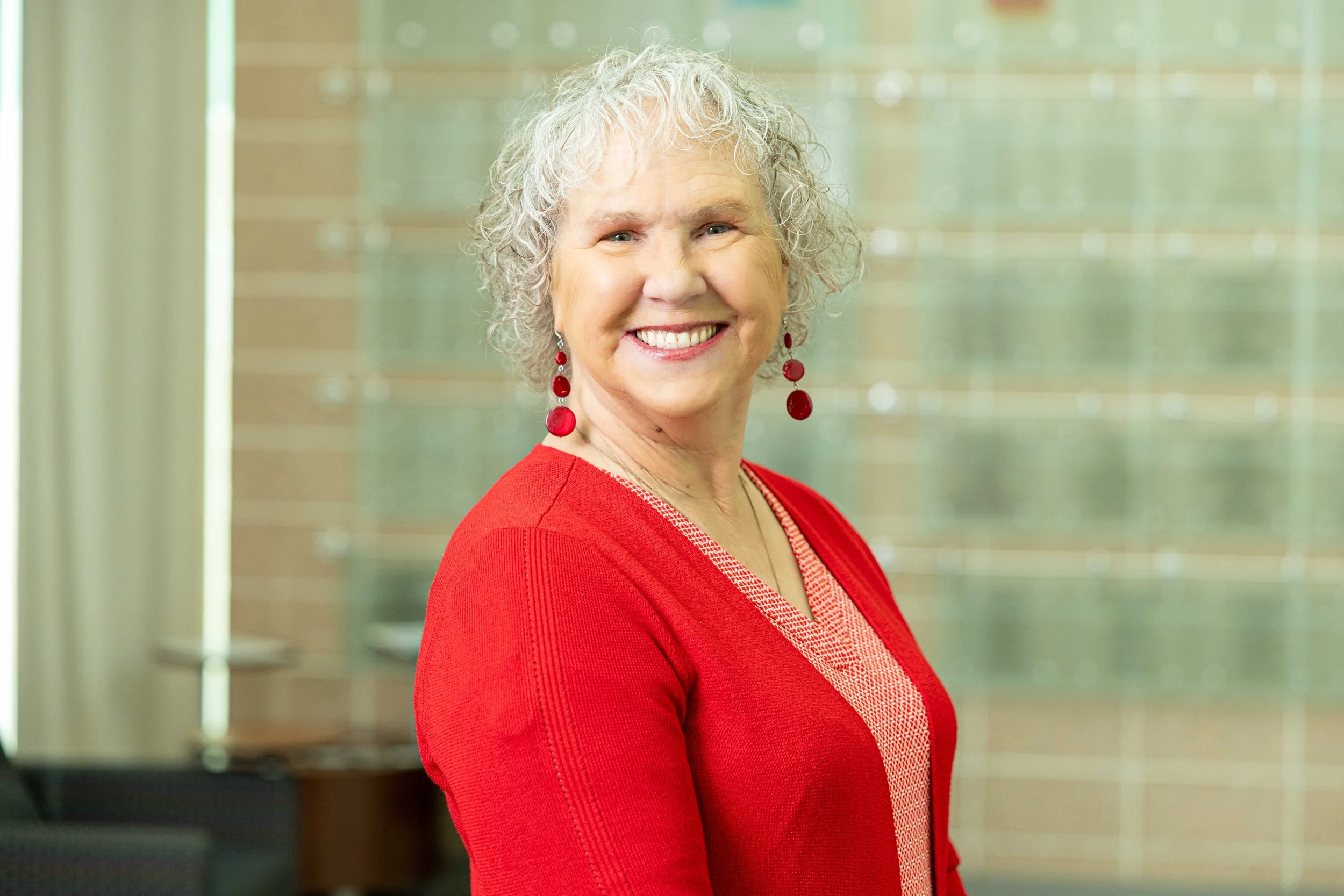 An older woman with curly gray hair, wearing a red cardigan and red earrings, smiling confidently in an indoor setting.
