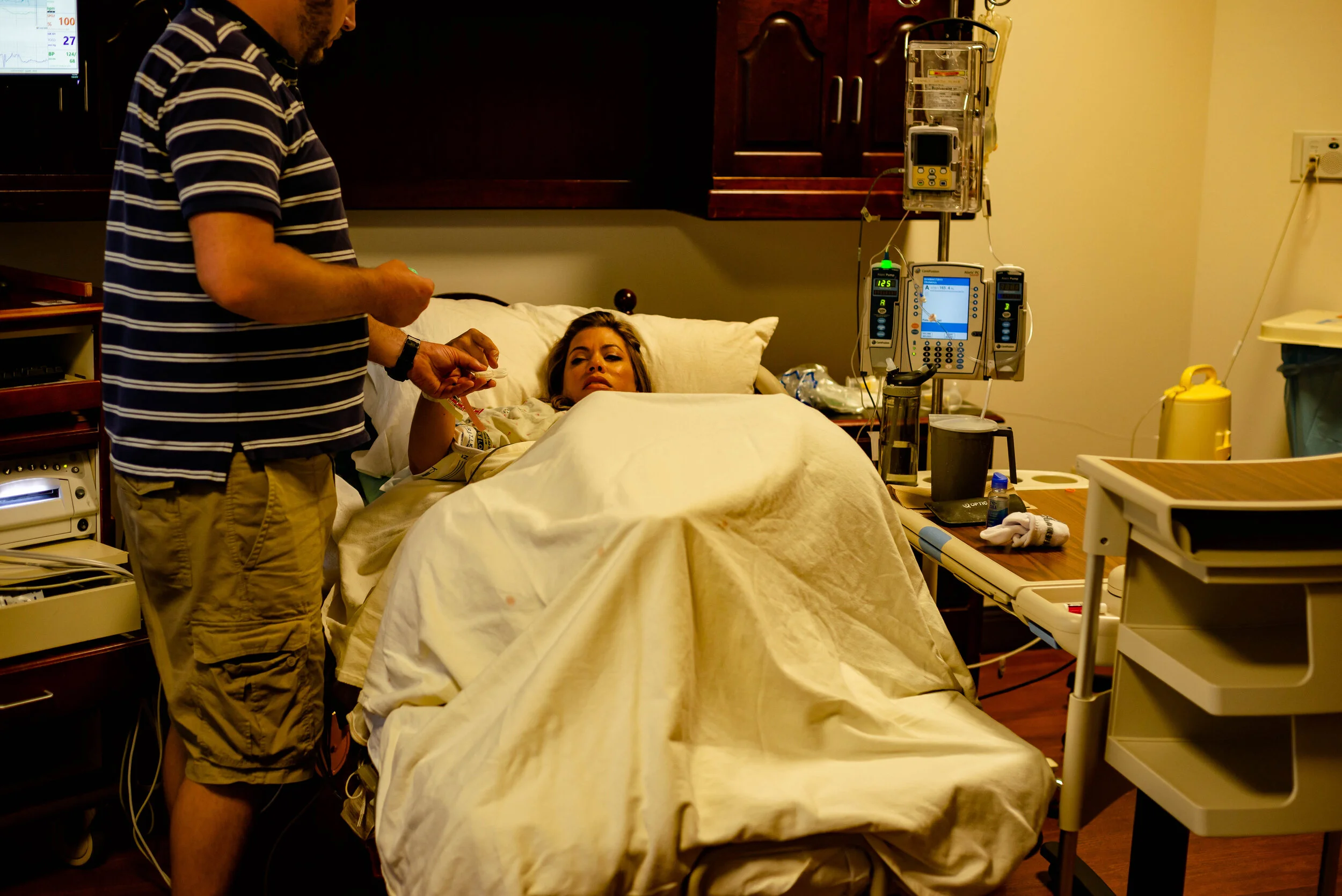 mom laying in hospital bed, removing contact lenses while her husband holds the contact lens case for her.