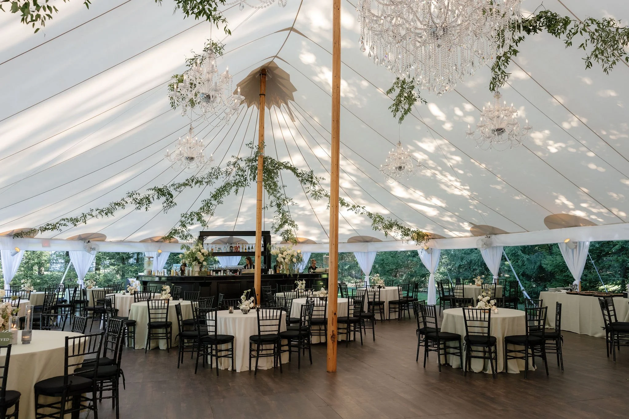 Elegant outdoor event tent decorated with chandeliers, greenery, and white curtains, with round tables set with white tablecloths and floral centerpieces, surrounded by black chairs.
