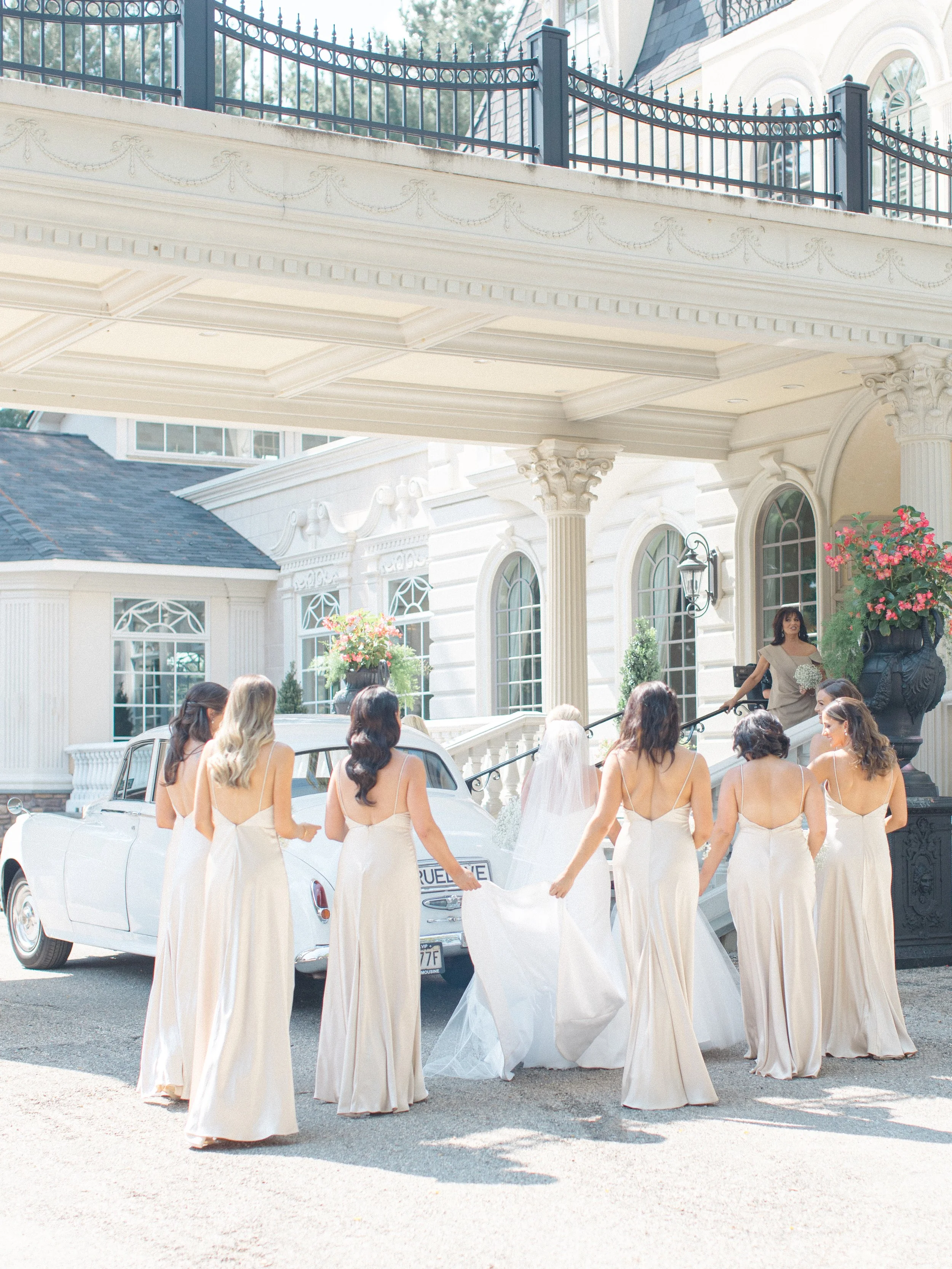 Bridesmaids in cream-colored dresses walk towards a bride under an ornate building entrance with columns, a staircase, and large flower arrangements.