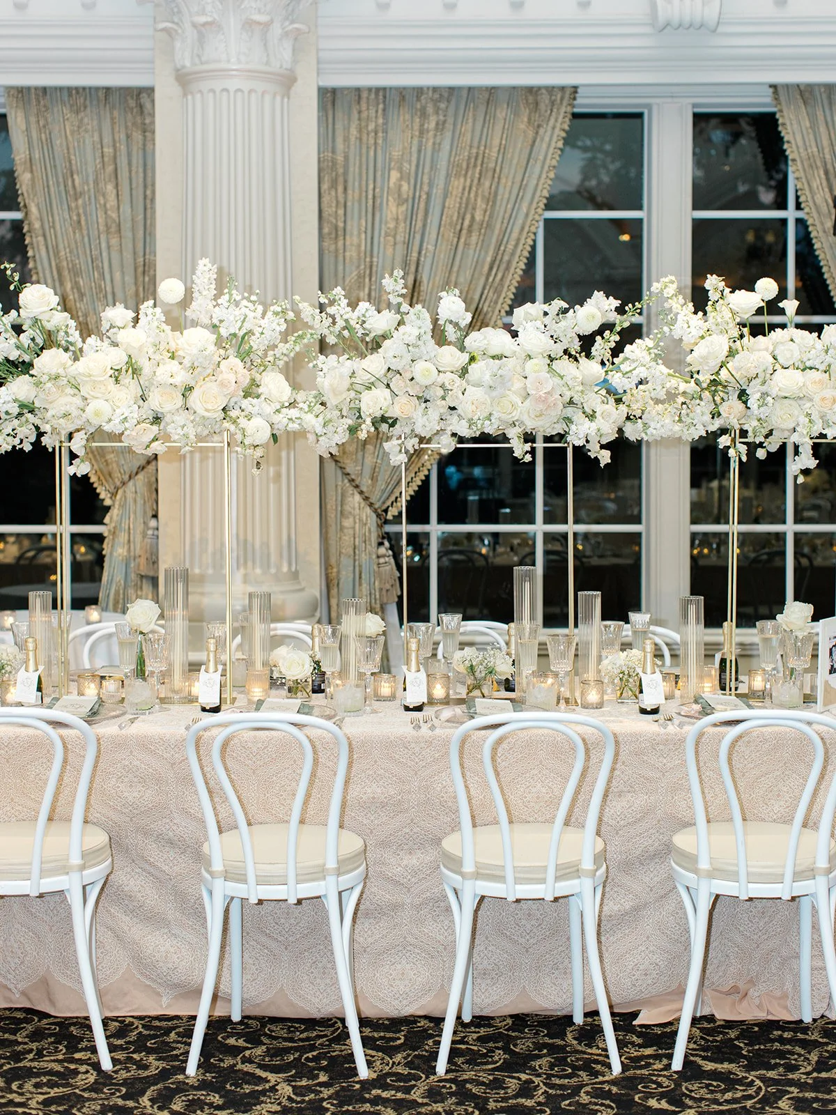 Elegant wedding reception table with white floral arrangements on tall gold stands, surrounded by white chairs, candles, and glassware, set in a luxurious room with draped curtains and large windows.