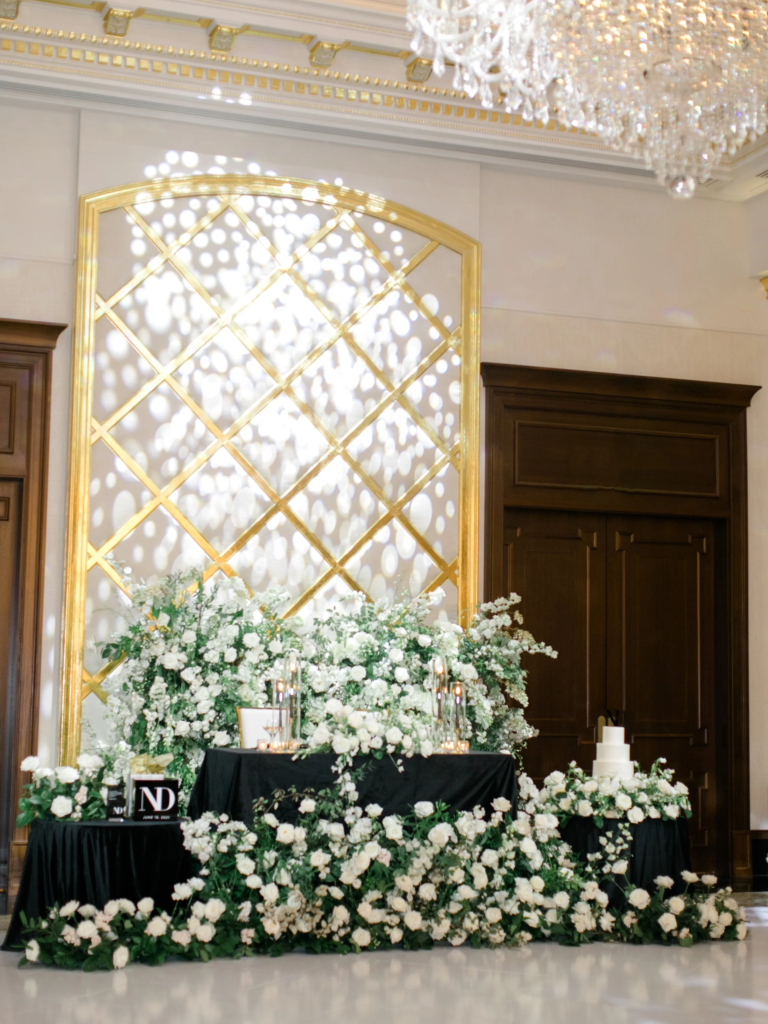 A wedding or event display with a backdrop of gold lattice and hanging light reflections, a table adorned with white flowers, candles, and a three-tiered wedding cake, all in an elegant indoor setting.
