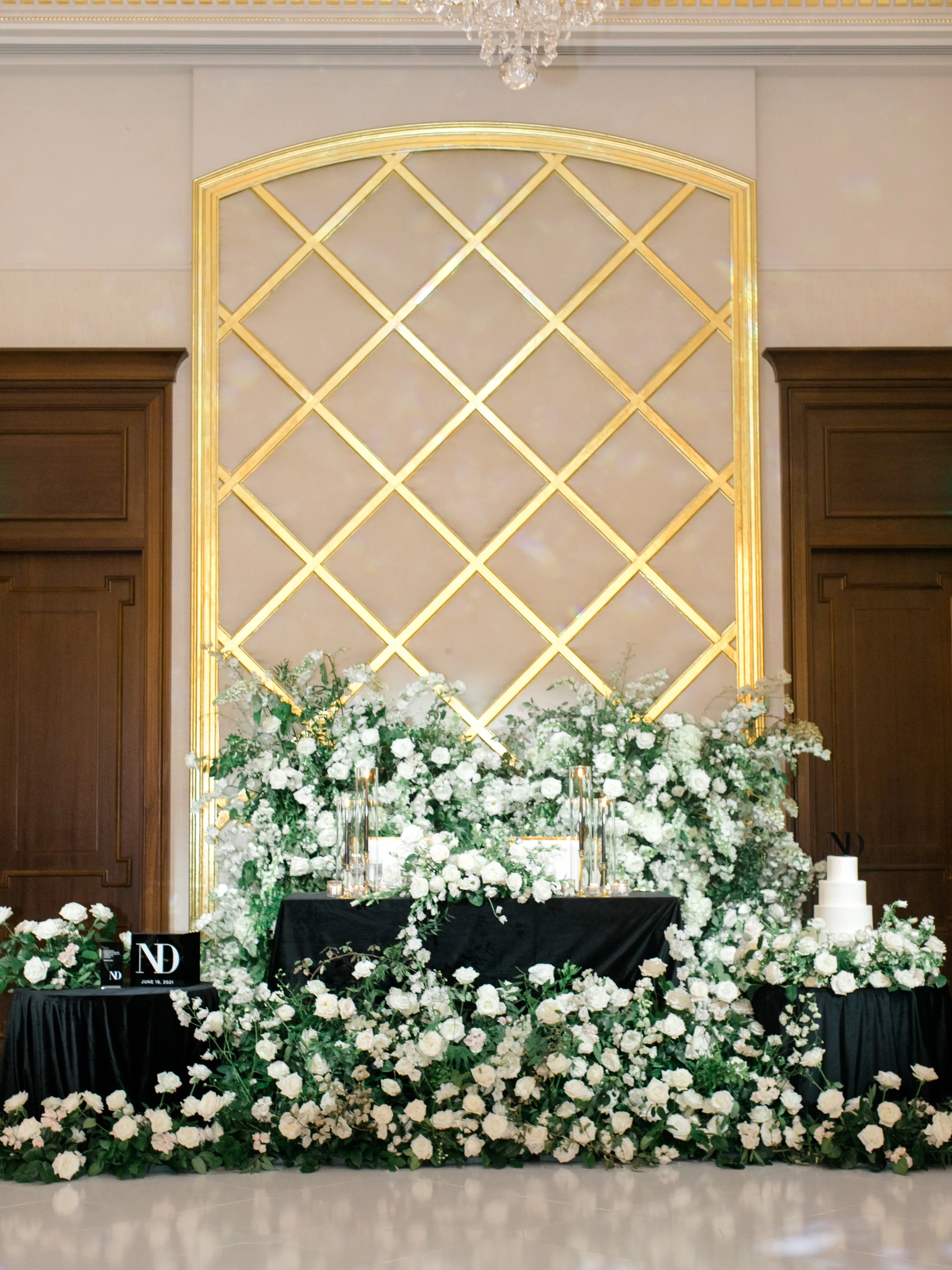 Wedding or event decor with white flowers, candles, and a cake, arranged on a black table in front of a gold geometric backdrop.