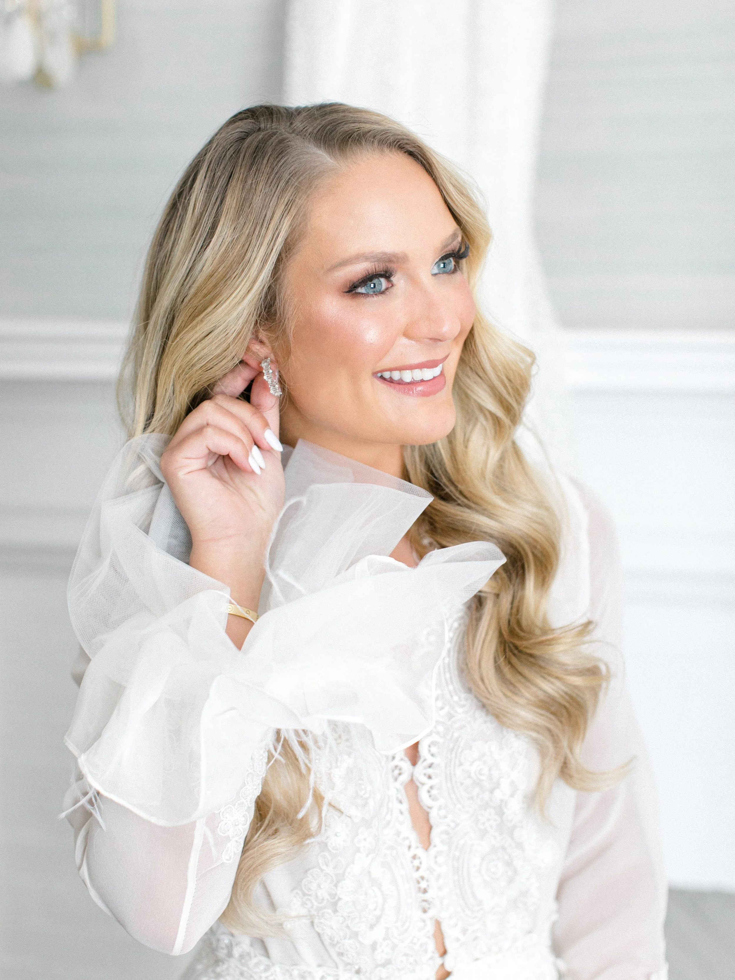 A woman with long blonde hair and bright blue eyes is smiling while holding an earring to her ear. She is wearing a white lace dress with voluminous, sheer ruffled sleeves and a high neckline. The background shows a white room with some elaborate dec