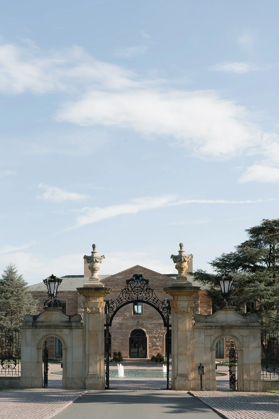 View of an elegant gated estate with stone pillars, decorative iron gate, fountains, and a historic-style building in the background, under a partly cloudy sky.