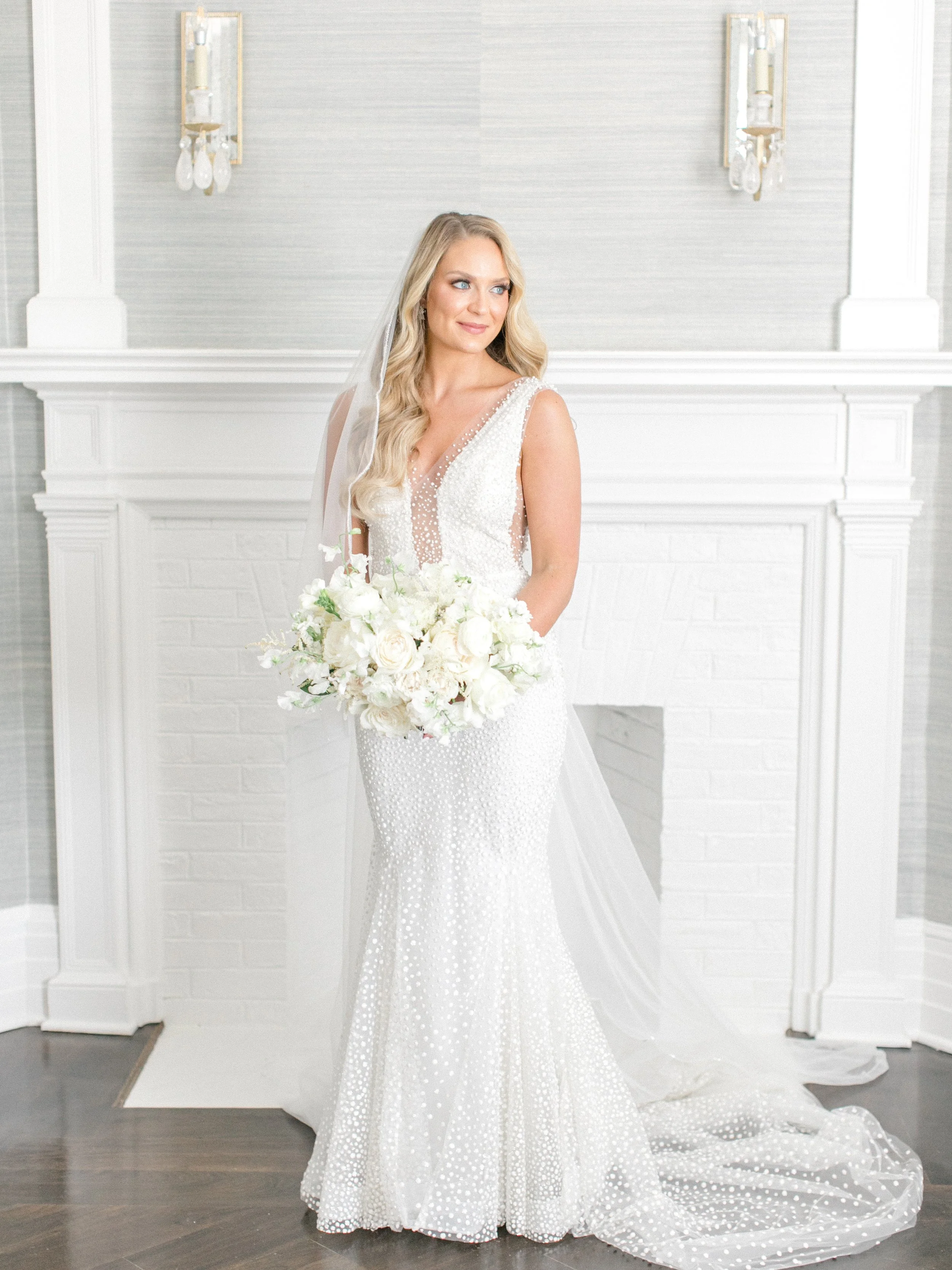 Bridal portrait of a bride in a white wedding gown holding a large bouquet of white flowers, standing in front of a white fireplace and wall with two light fixtures.