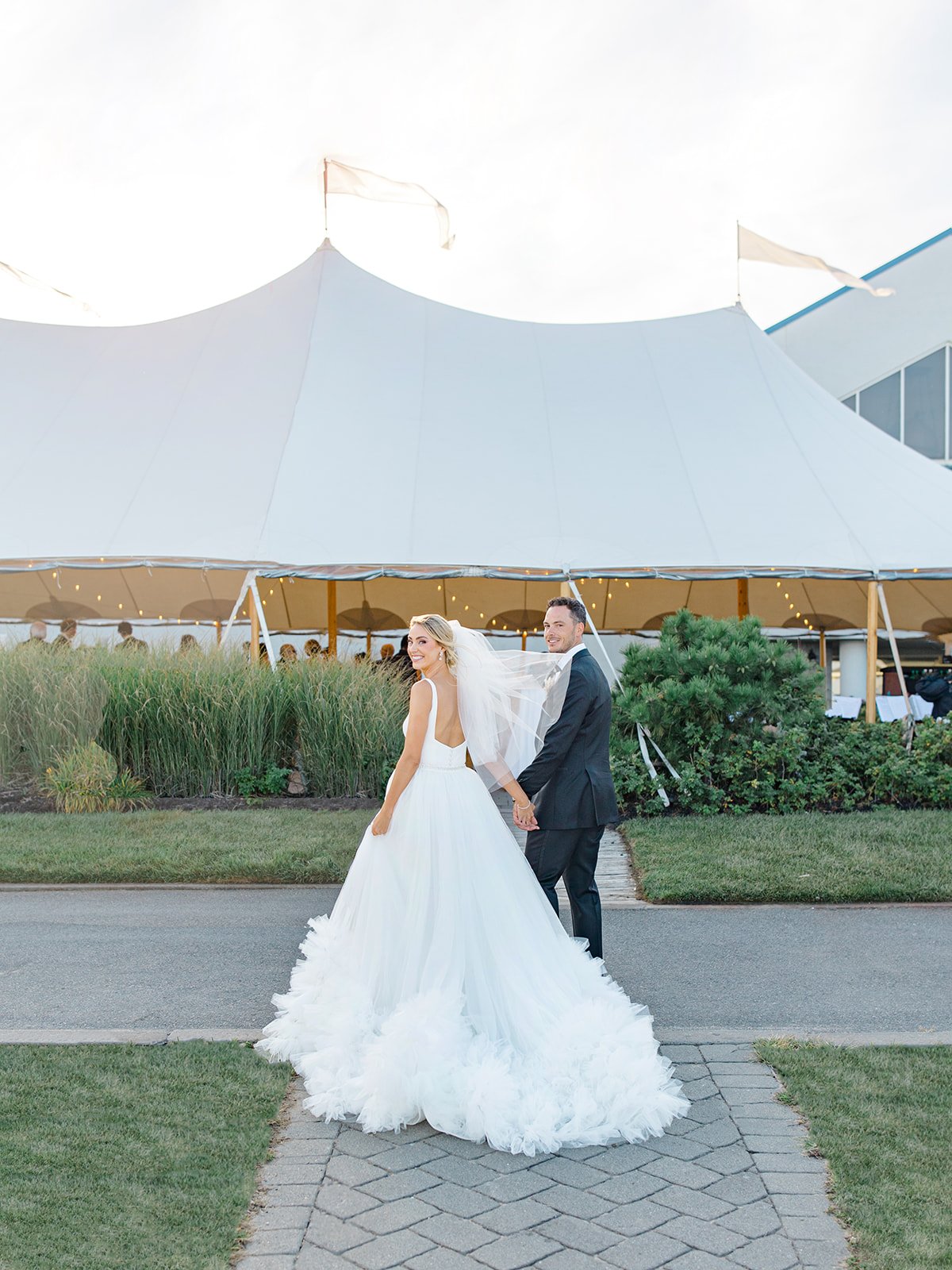 Bride and groom holding hands outside wedding tent, bride in a white wedding gown and veil, groom in a dark suit, with tent and string lights in background.