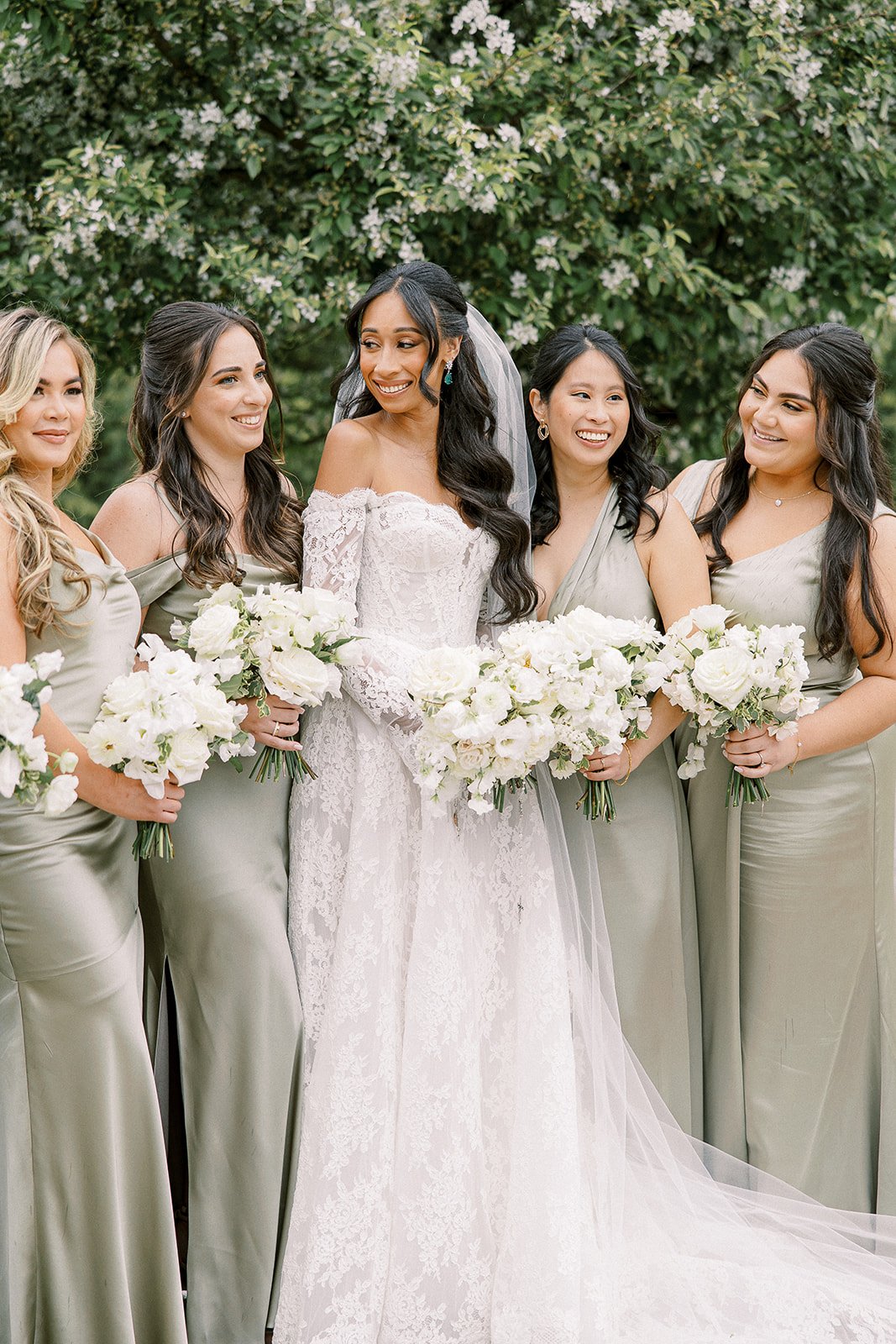 A bride with dark wavy hair in a white lace wedding dress and veil, standing among five bridesmaids in light sage green satin dresses, holding white floral bouquets outdoors with green foliage and white flowers in the background.