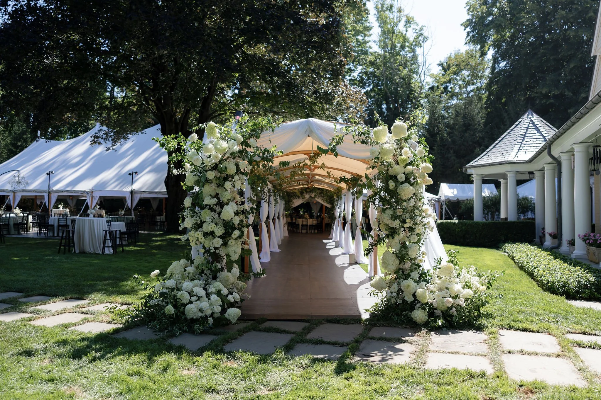 A decorated outdoor wedding ceremony arch with white flowers and greenery, surrounded by a lush garden, tents with tables and chairs in the background, and a clear sunny sky.