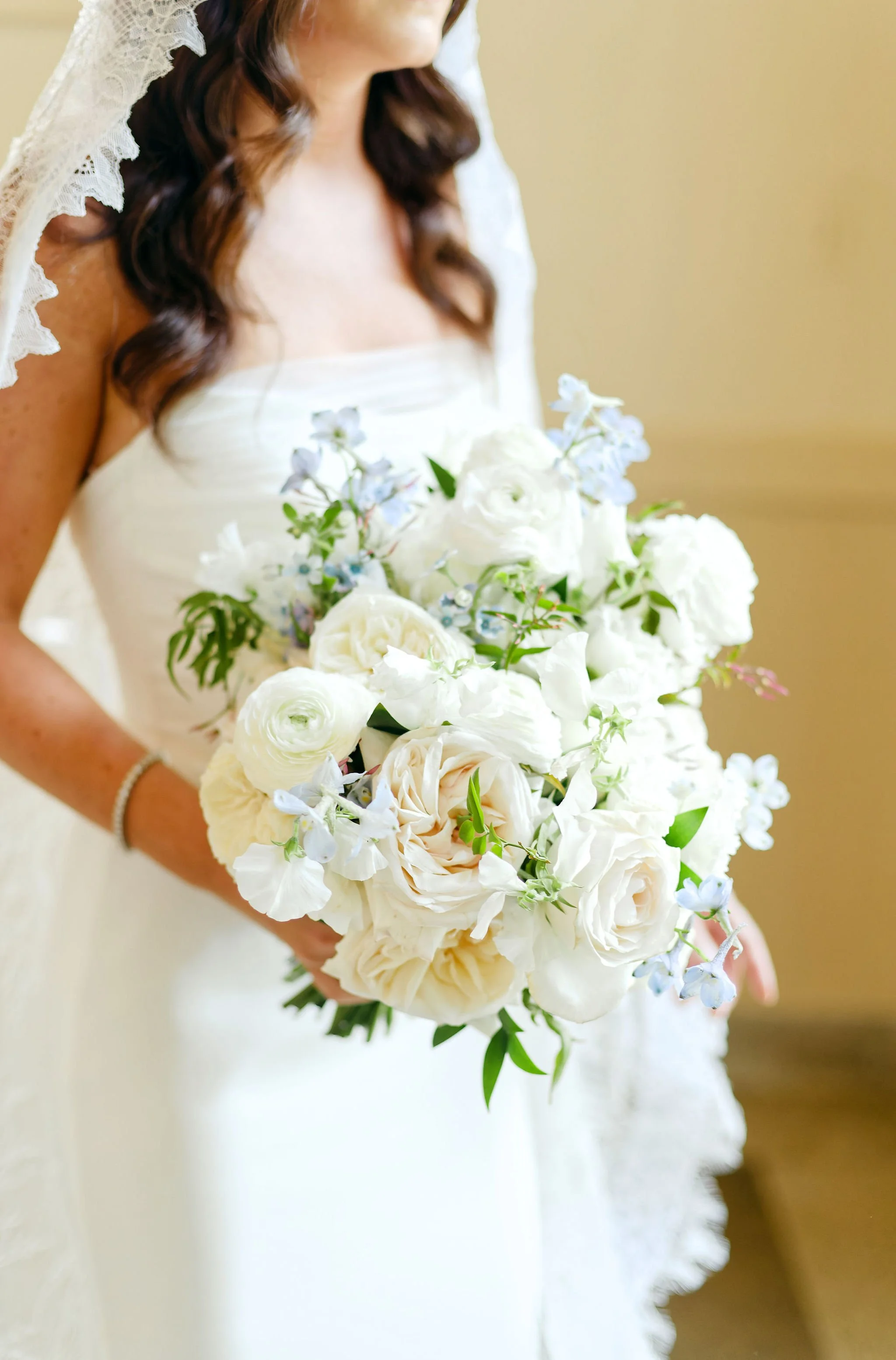 Close-up of a bride in a strapless wedding dress holding a bouquet of white flowers, including roses and other blooms, with her face partially out of frame.