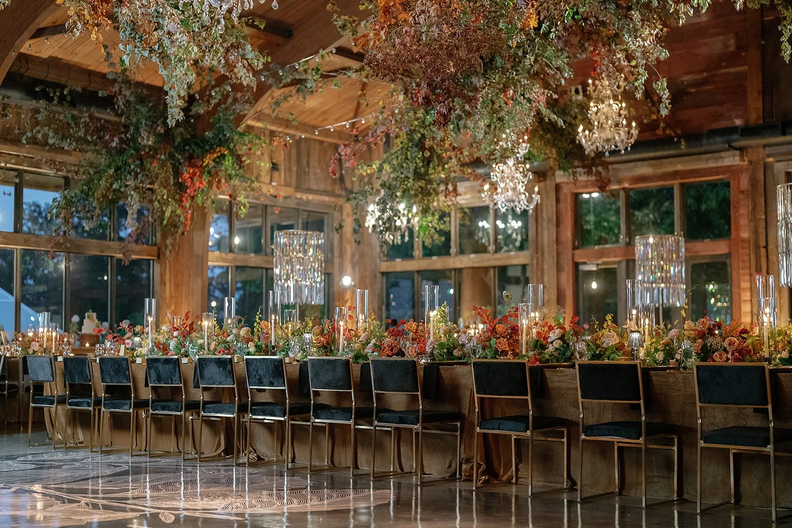 Elegant banquet table decorated with flowers, candles, and glassware in a rustic wooden hall with large windows, chandeliers, and greenery overhead.