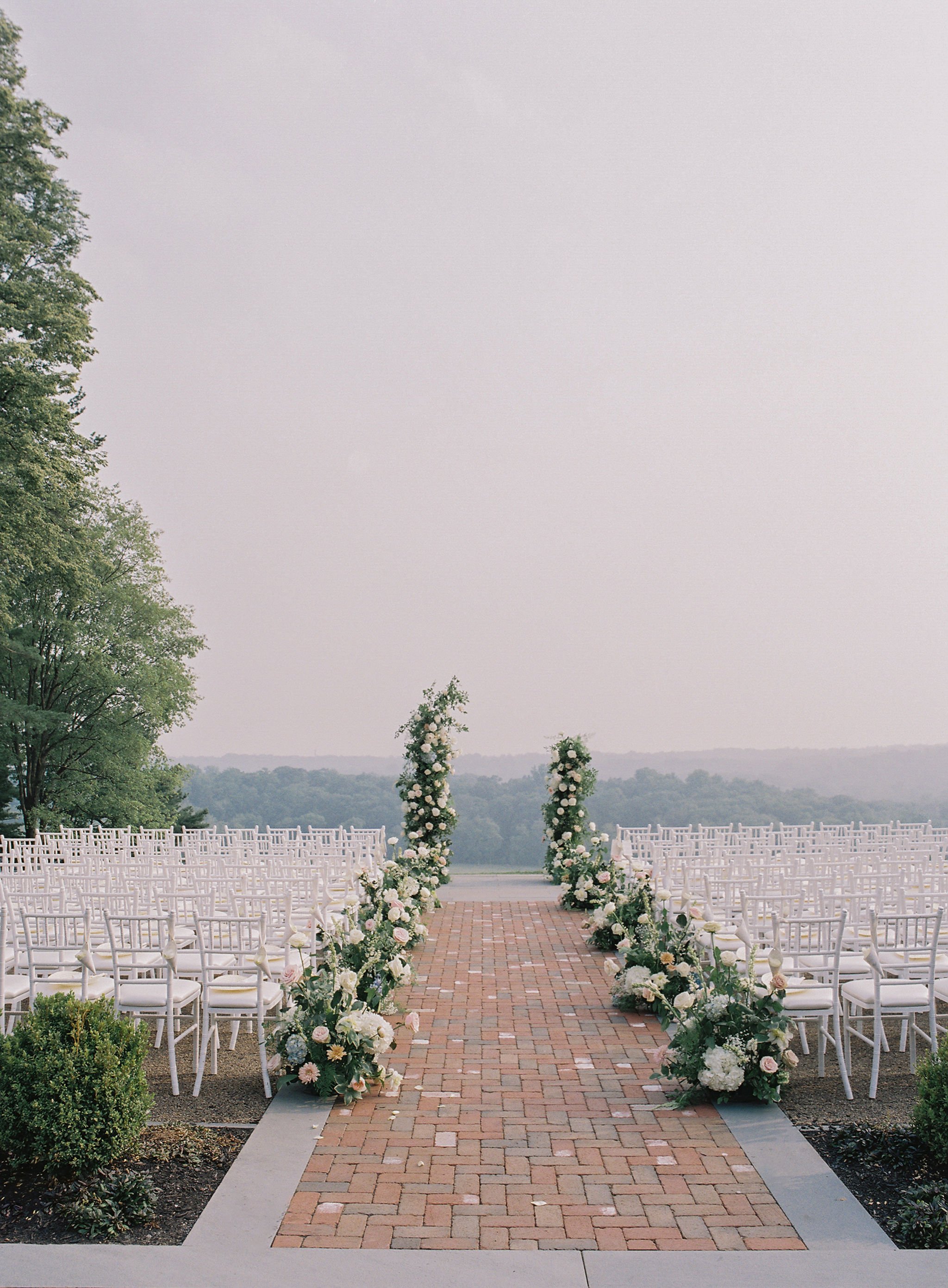 Outdoor wedding ceremony setup with rows of white chairs on either side of a brick aisle, decorated with floral arrangements of white, pink, and green flowers.
