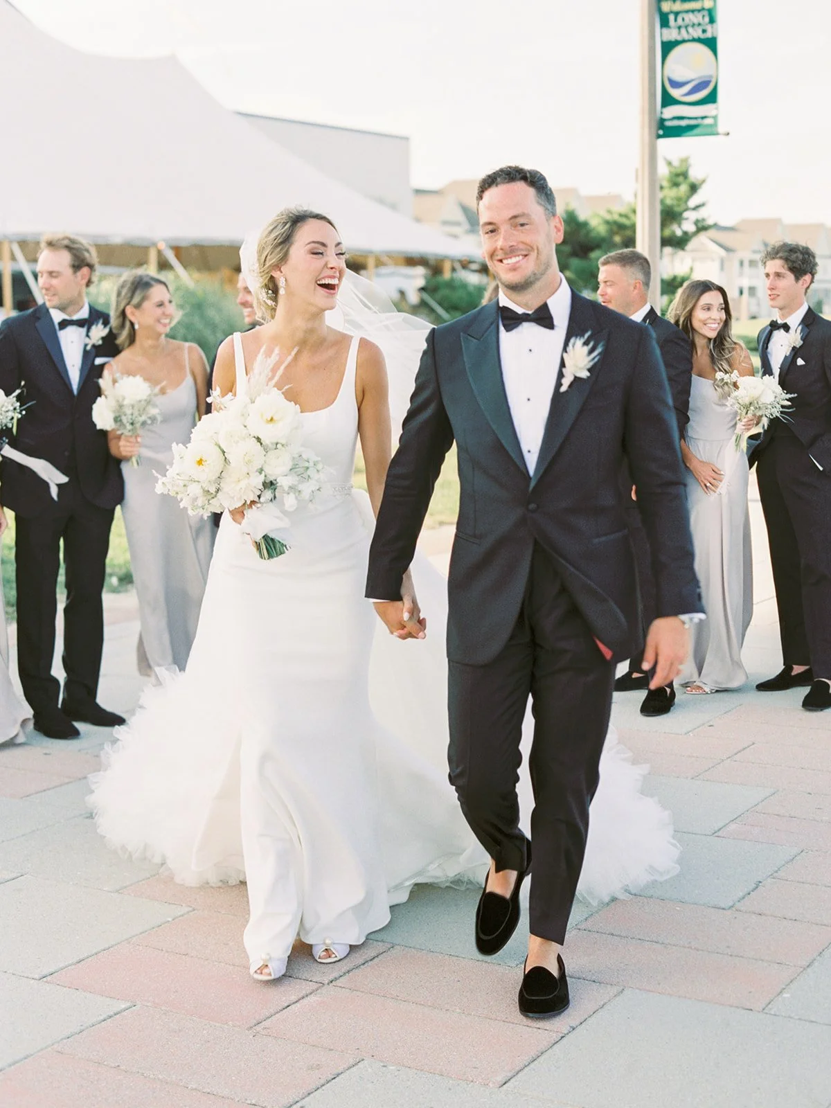 A bride and groom walking hand in hand, smiling, surrounded by wedding guests in formal attire at an outdoor wedding.