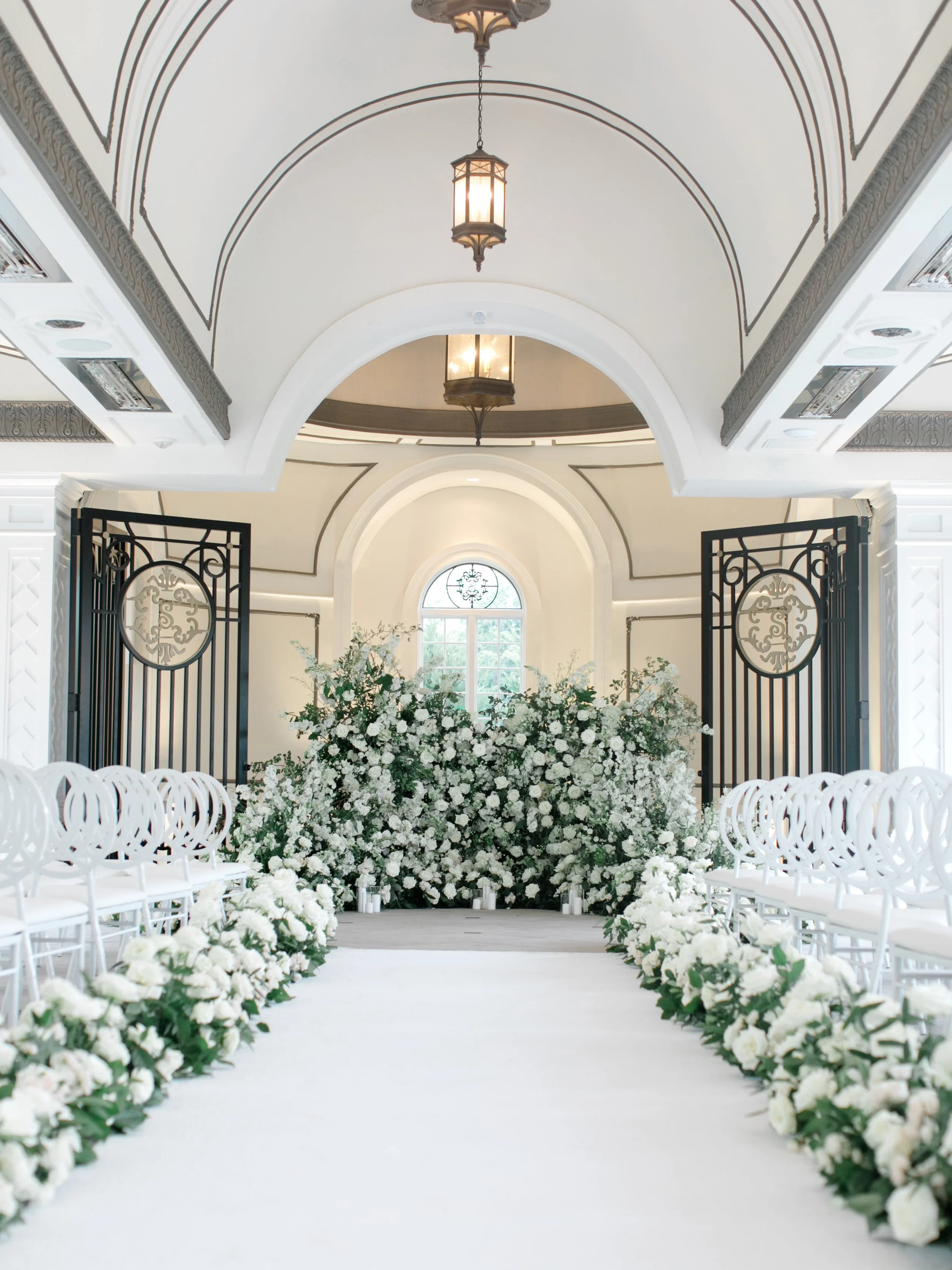 Elegant wedding ceremony aisle decorated with white flowers and greenery leading to an altar with floral arrangements and a window in the background, inside a bright, ornate hall.