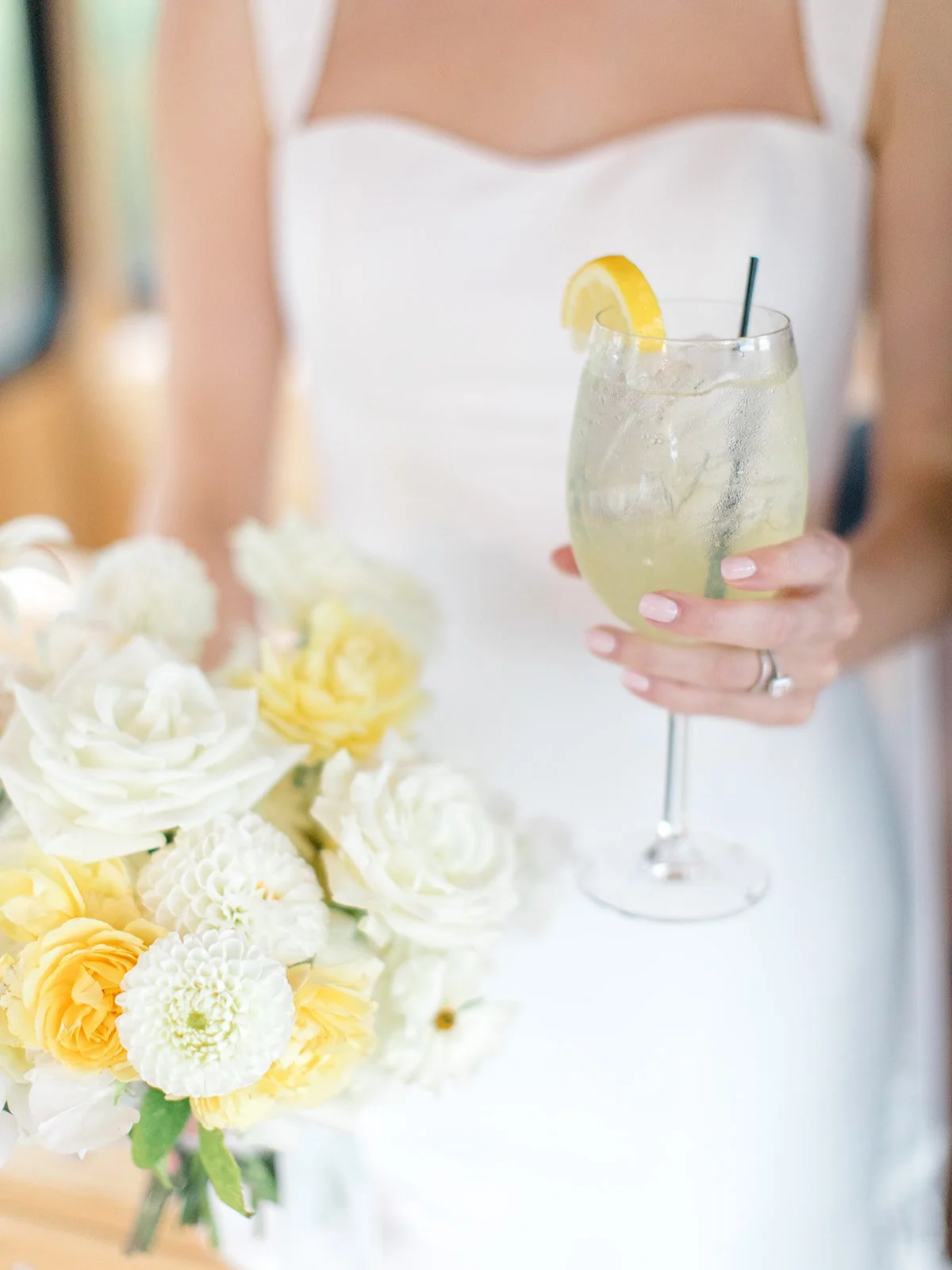 A person in a white dress holding a glass of lemonade with a lemon slice, standing next to a bouquet of white and yellow flowers.