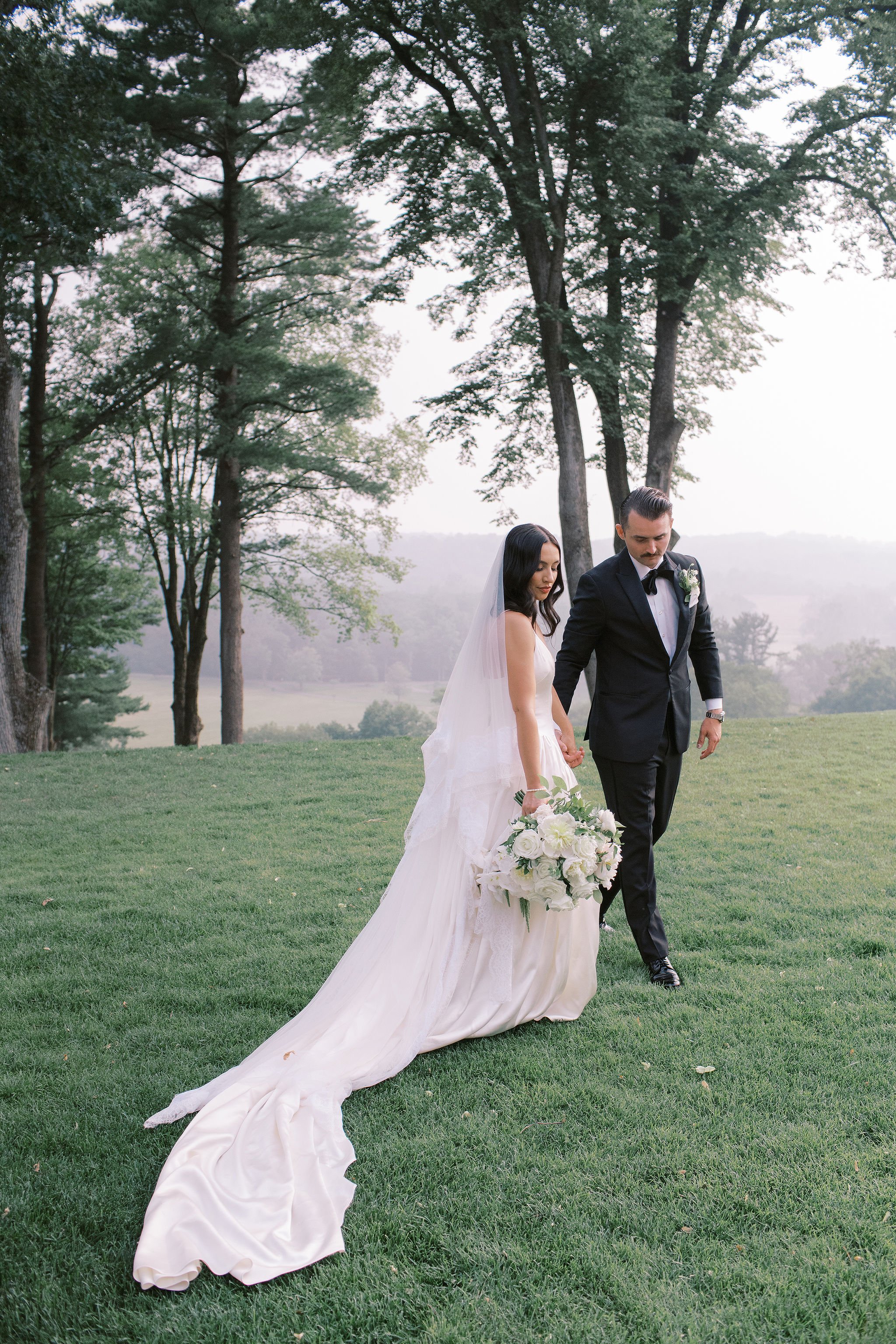 Bride and groom walking on grass in a park or garden during daytime, surrounded by trees, with the bride wearing a white wedding gown and veil, holding a bouquet of white flowers, and the groom dressed in a black tuxedo with a white shirt and bow tie.