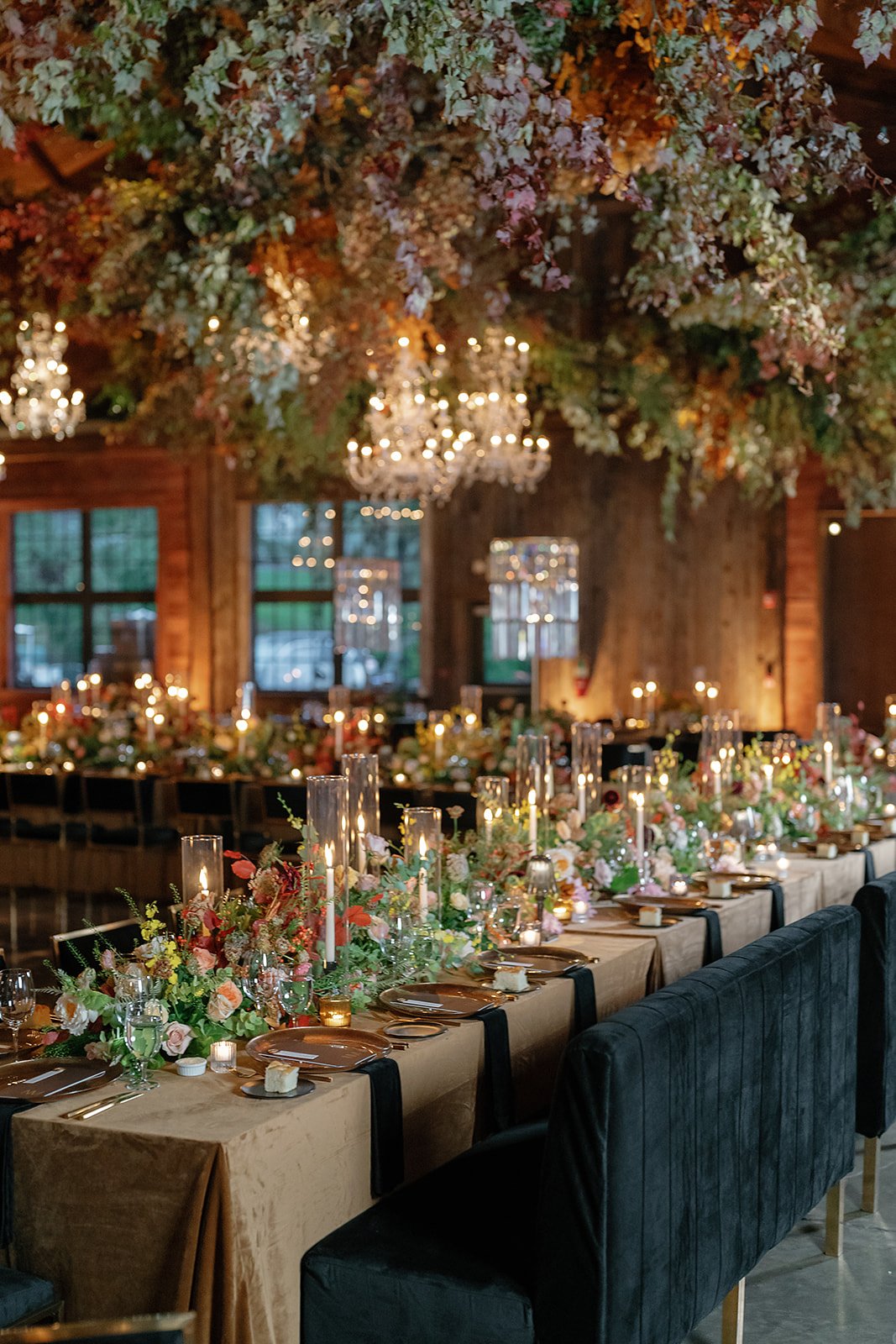 Elegant wedding reception with long tables decorated with floral arrangements and tall candles, chandeliers hanging from a ceiling with flowers, in a rustic wooden venue.