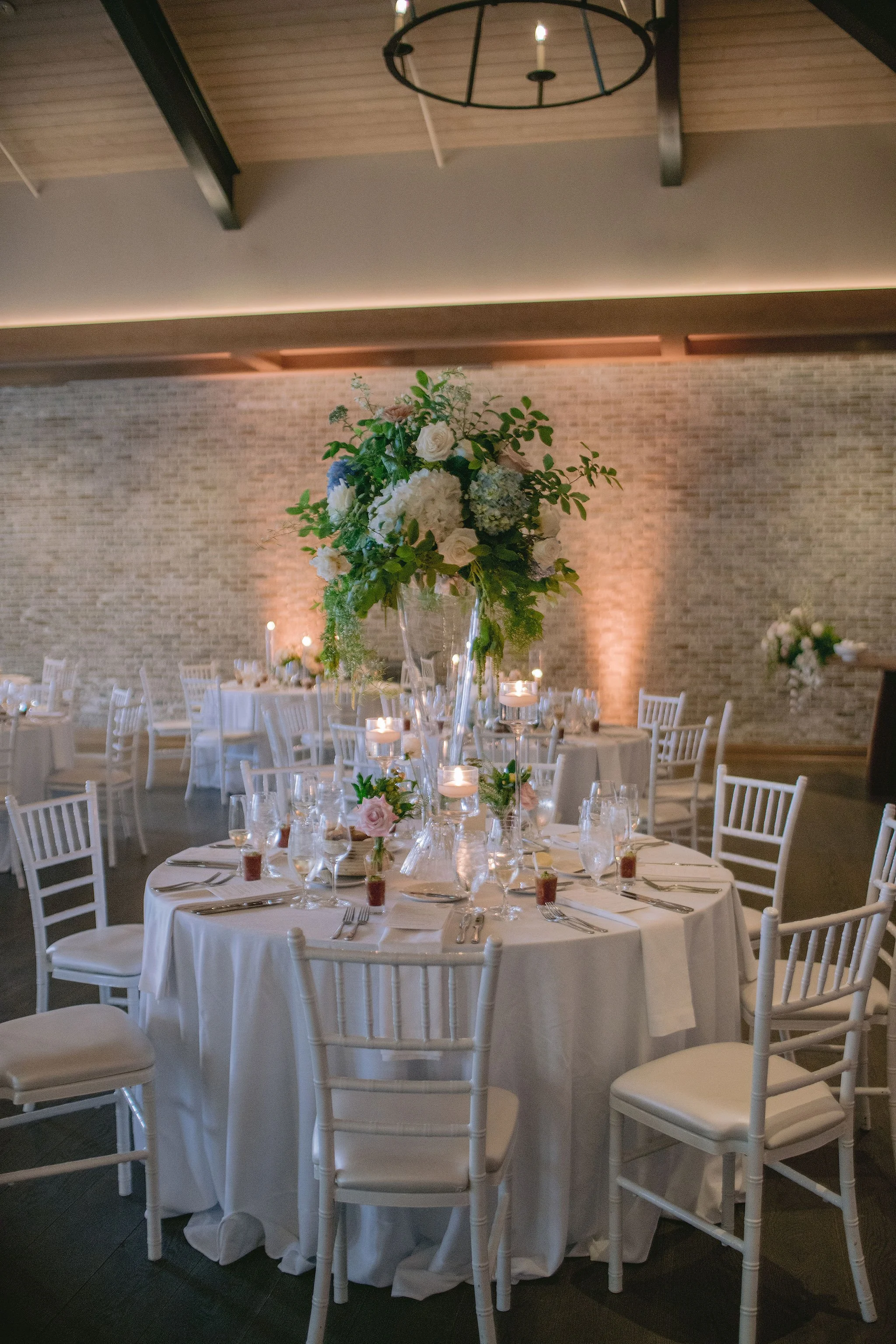 Wedding reception table with a tall floral centerpiece, set for dinner with candles, glassware, and silverware in a decorated banquet hall.