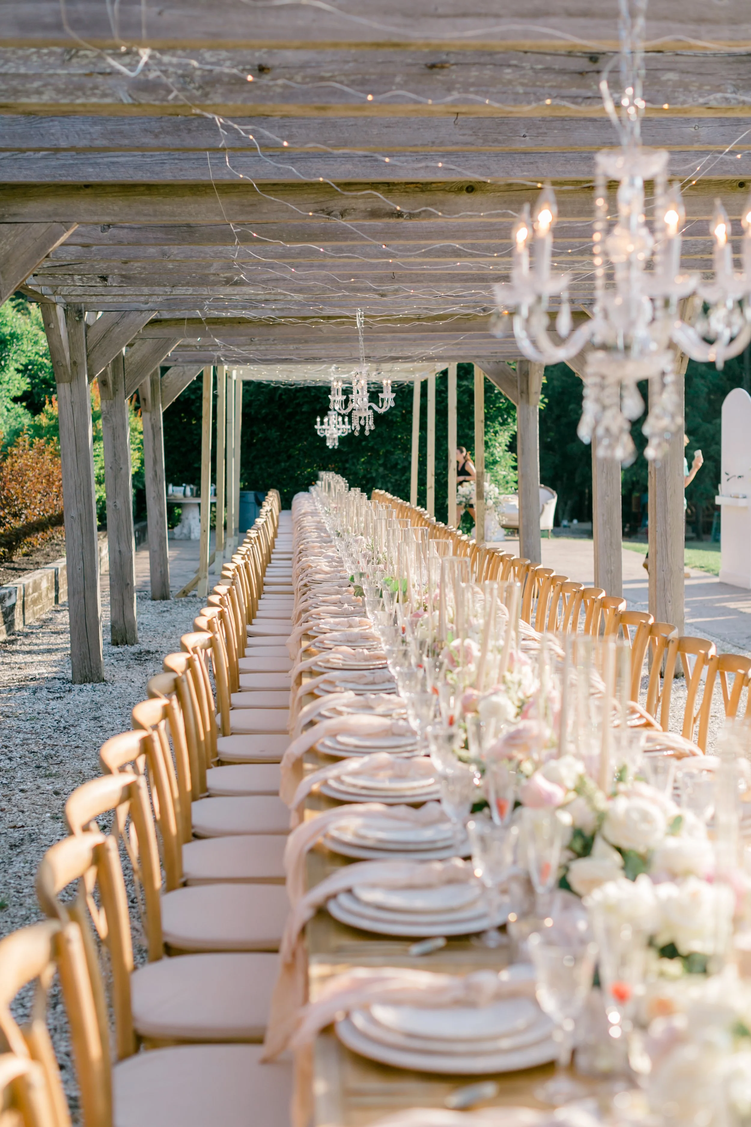 Long outdoor dining table decorated with white flowers and elegant tableware, under a rustic wooden canopy with chandeliers and string lights, set for a celebration or wedding reception.