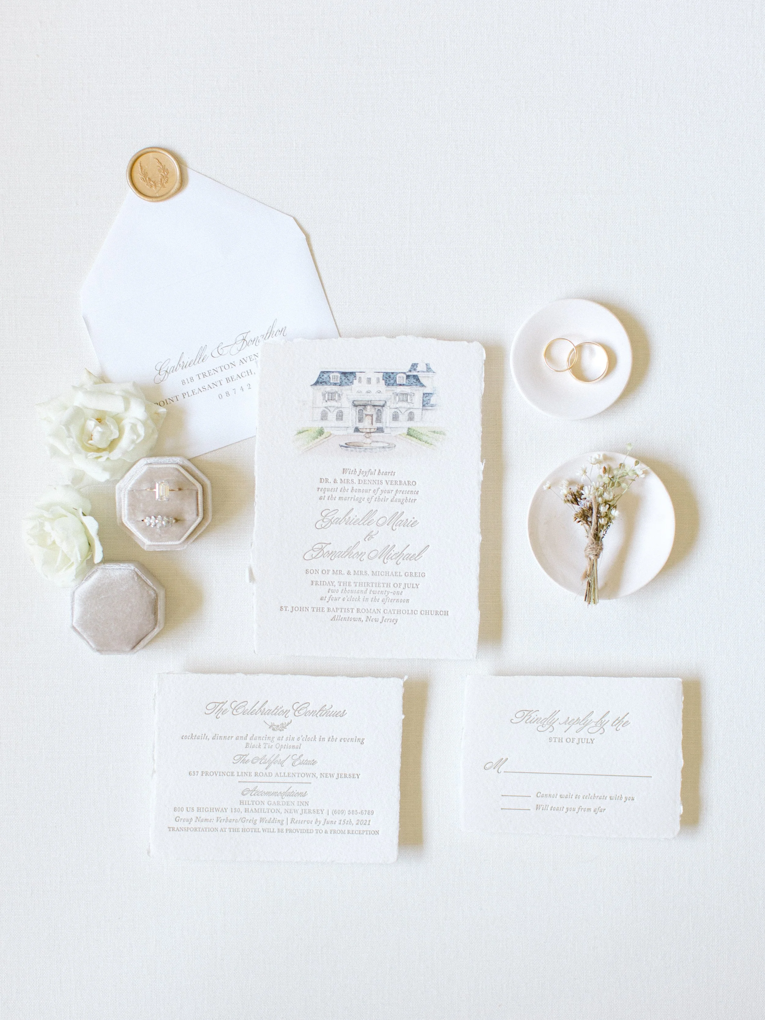 Wedding invitation set with floral accents, wedding rings, and small decorative items arranged on a white background.