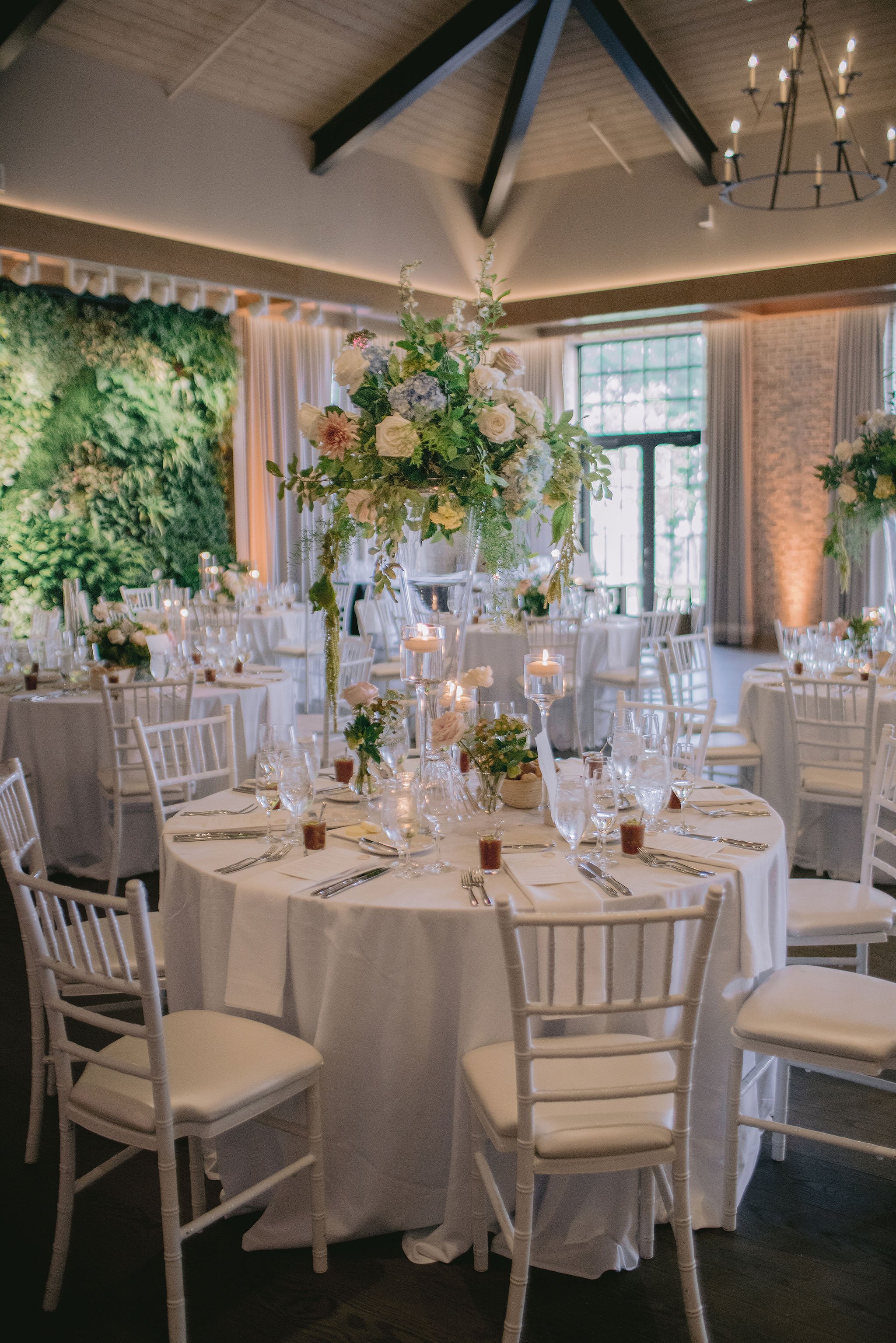 Wedding reception setup with round tables covered in white tablecloths, decorated with tall floral centerpieces, candles, and place settings, in a decorated hall with a green wall, curtains, and large windows.