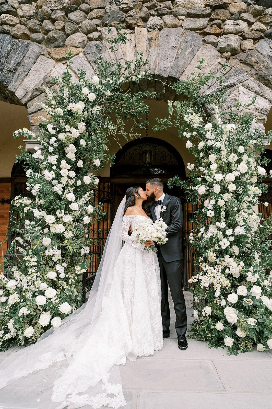 A bride and groom kissing under a floral archway at their wedding, with the bride in a white lace gown and the groom in a black tuxedo, outside the building with stone and brick walls.