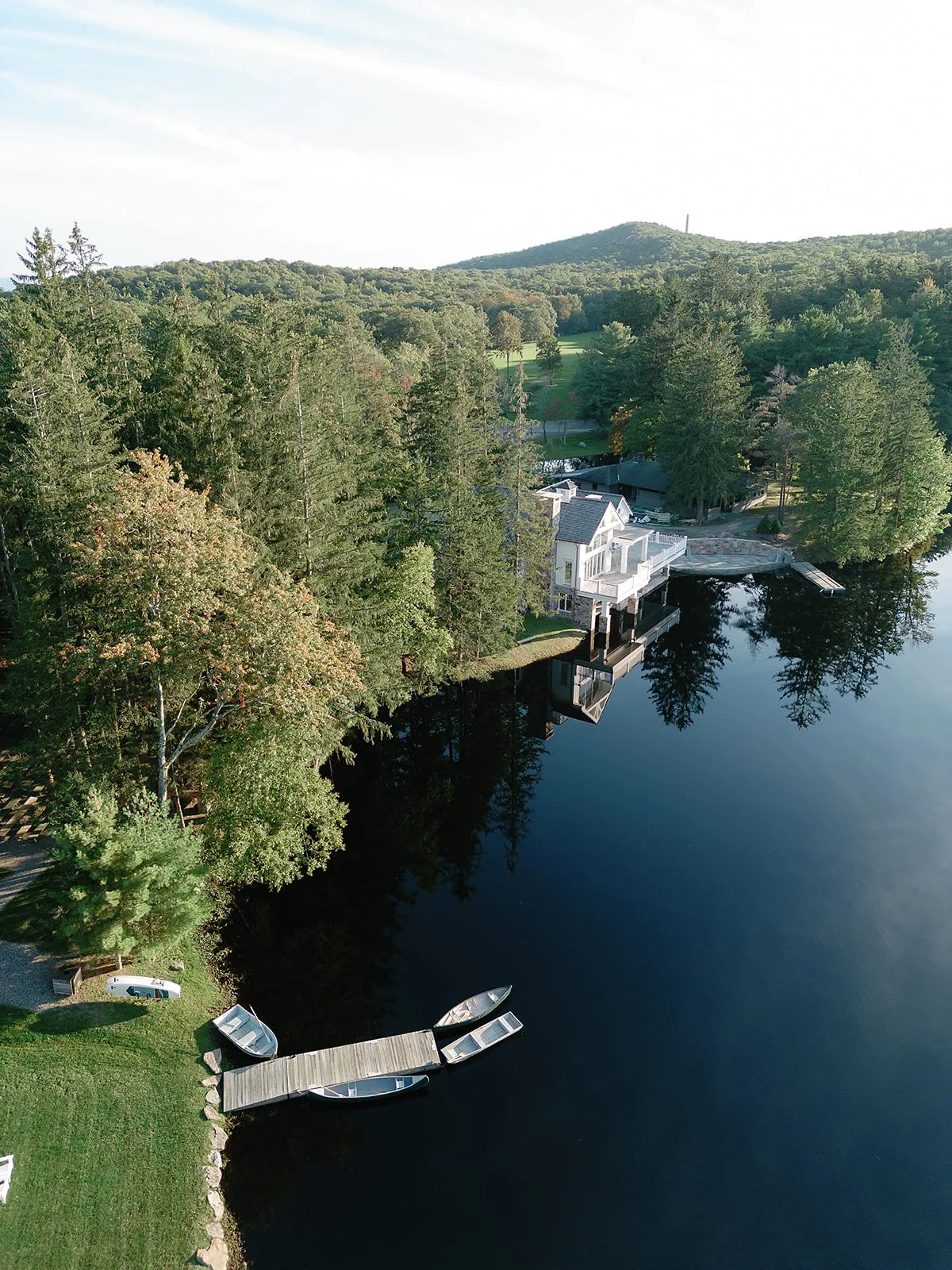 Aerial view of a lakefront property with a modern white house, dock, small boats, surrounded by trees and distant hills.